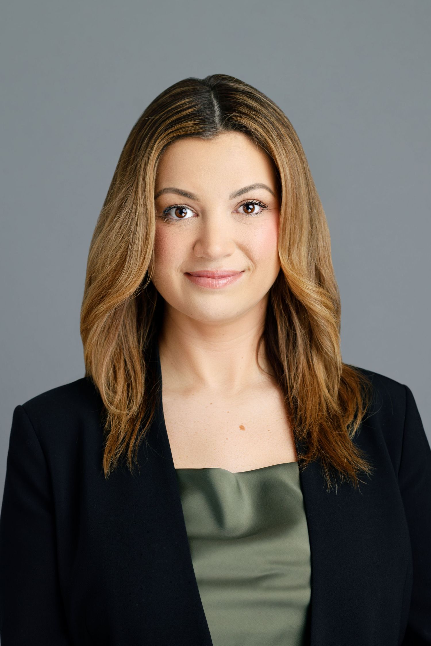 Professional headshot of a woman with long brown hair wearing a black blazer and green top against a gray background.