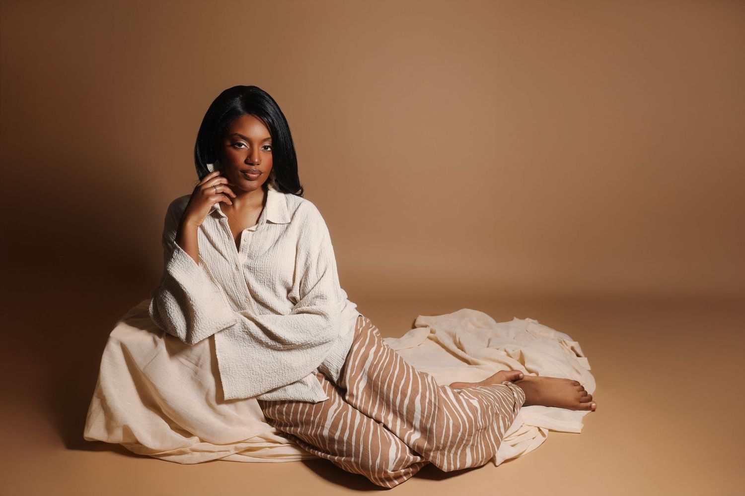 Woman in cream oversized shirt and striped pants sitting gracefully on beige studio floor.