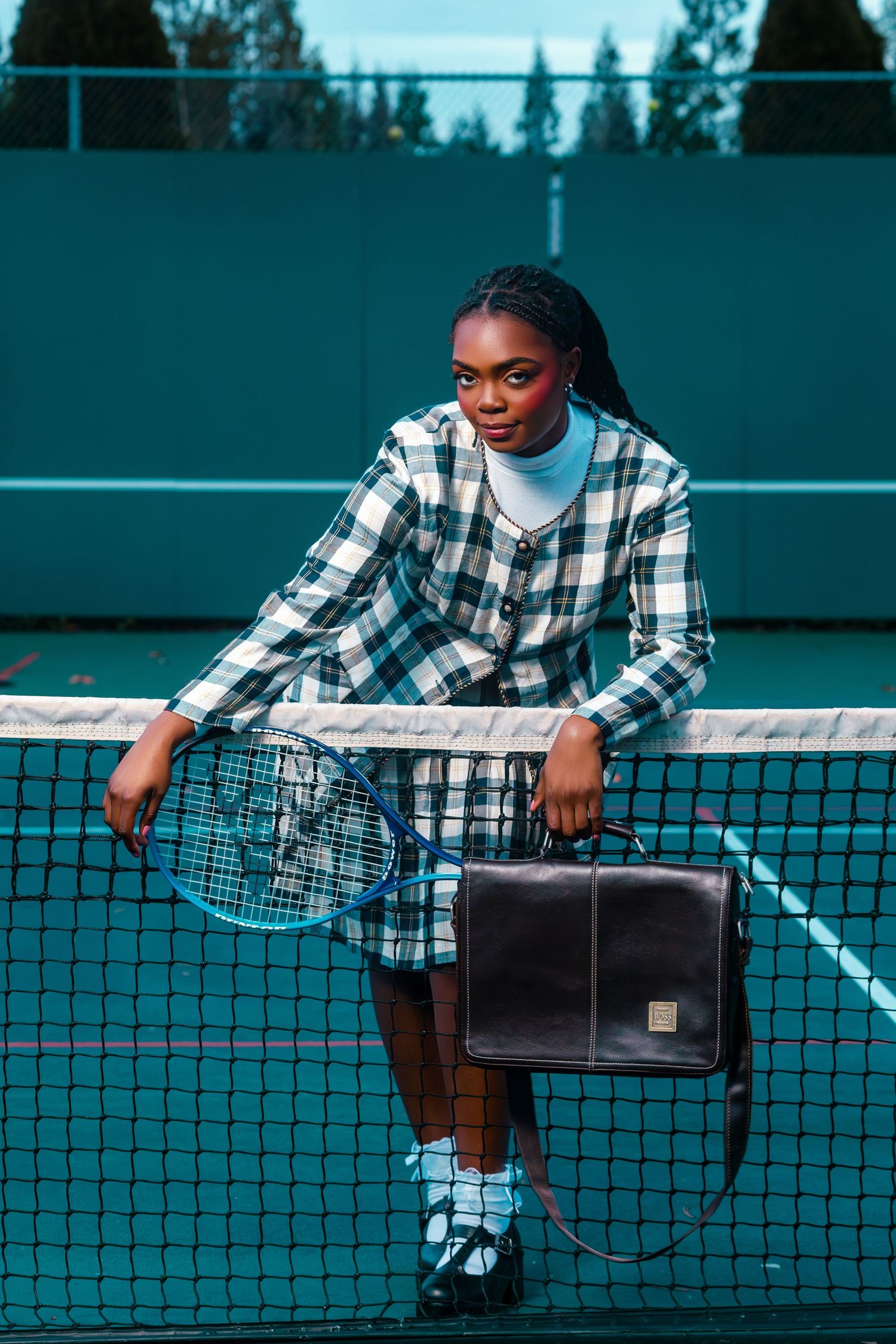 Young Black woman in plaid suit holds tennis racket and briefcase leaning on net at tennis court.