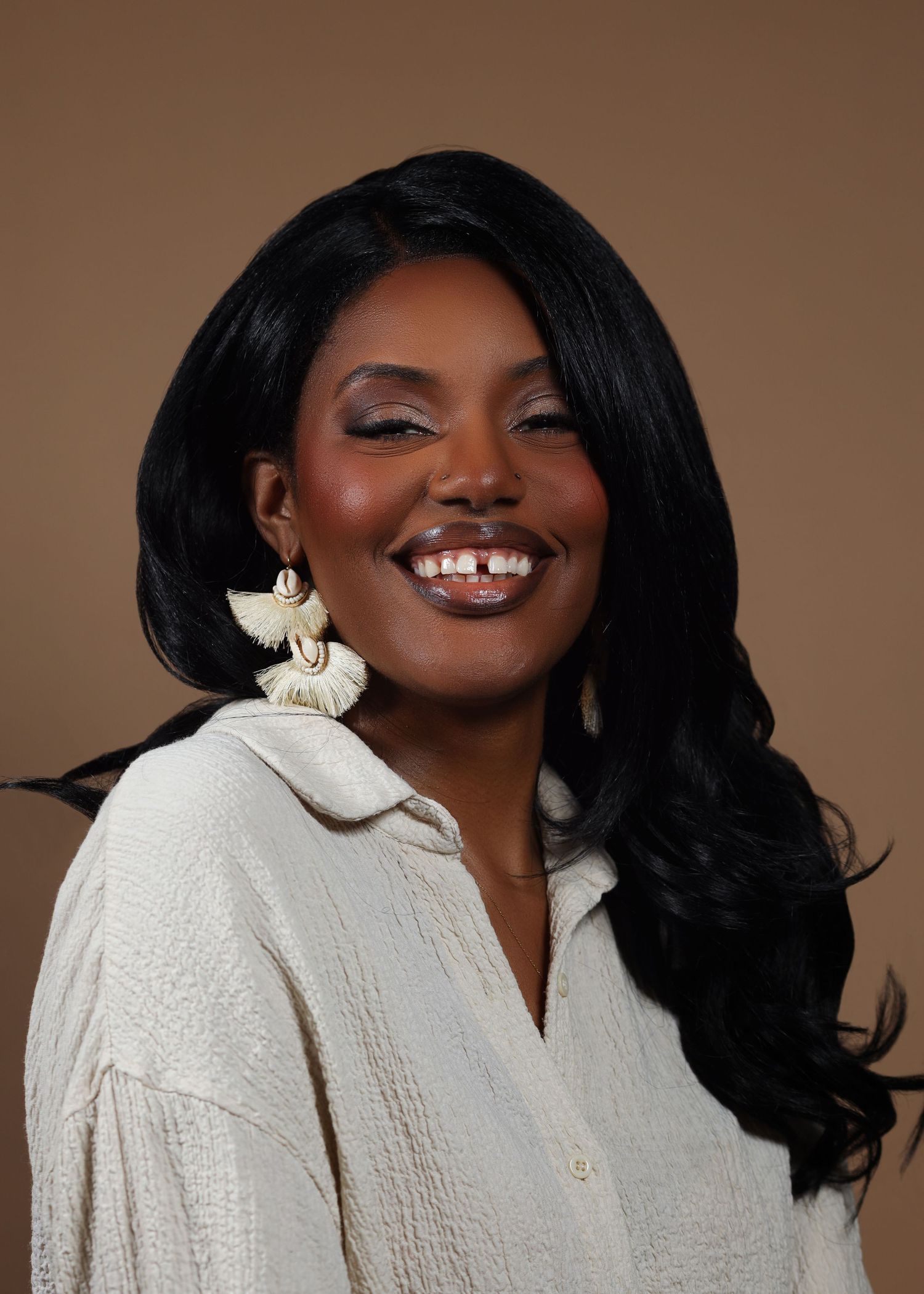 Smiling Black woman with long wavy hair wearing white tassel earrings and a white textured blouse against a tan background.