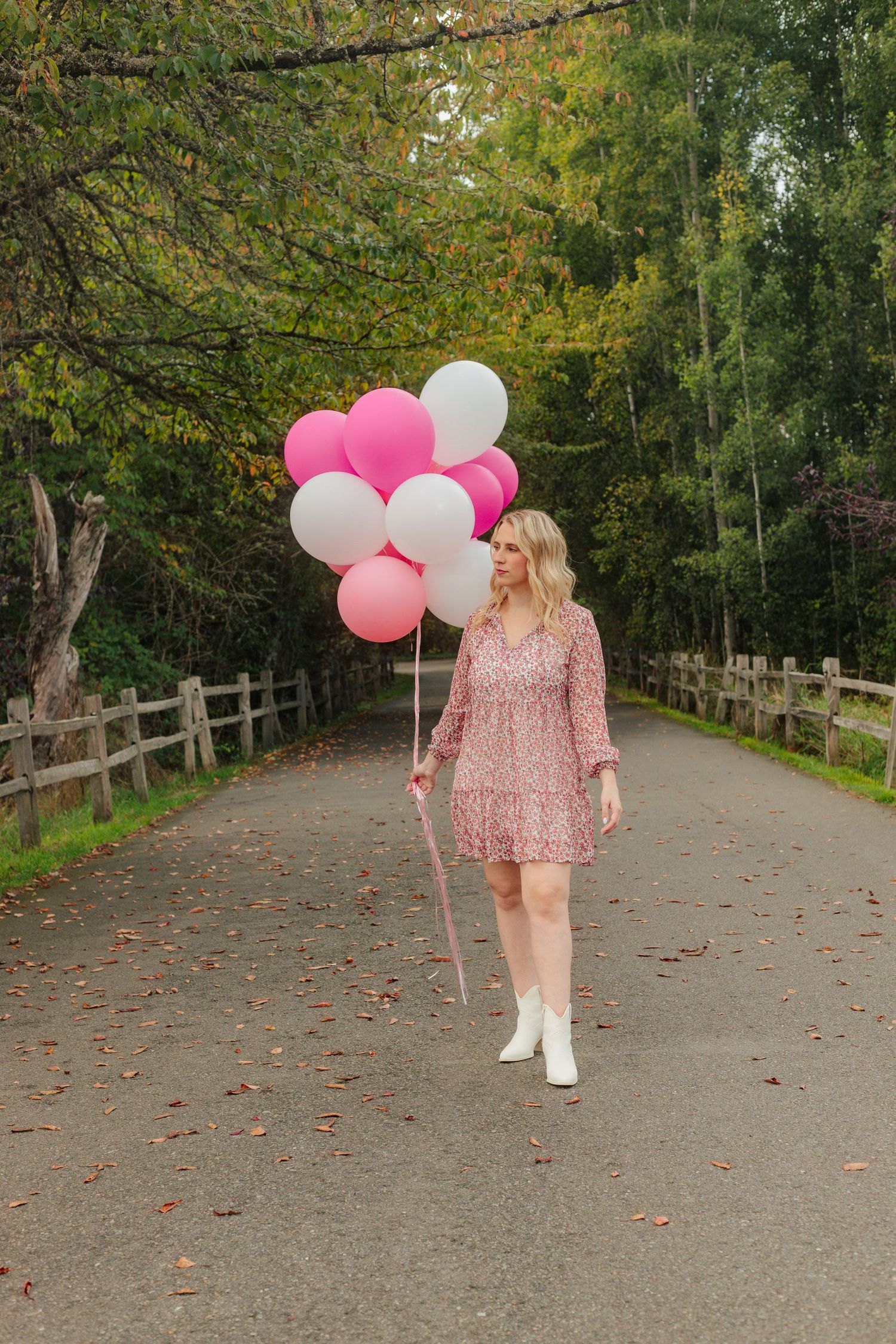 Person walking down a tree-lined path with pink and white balloons and wearing a floral dress.