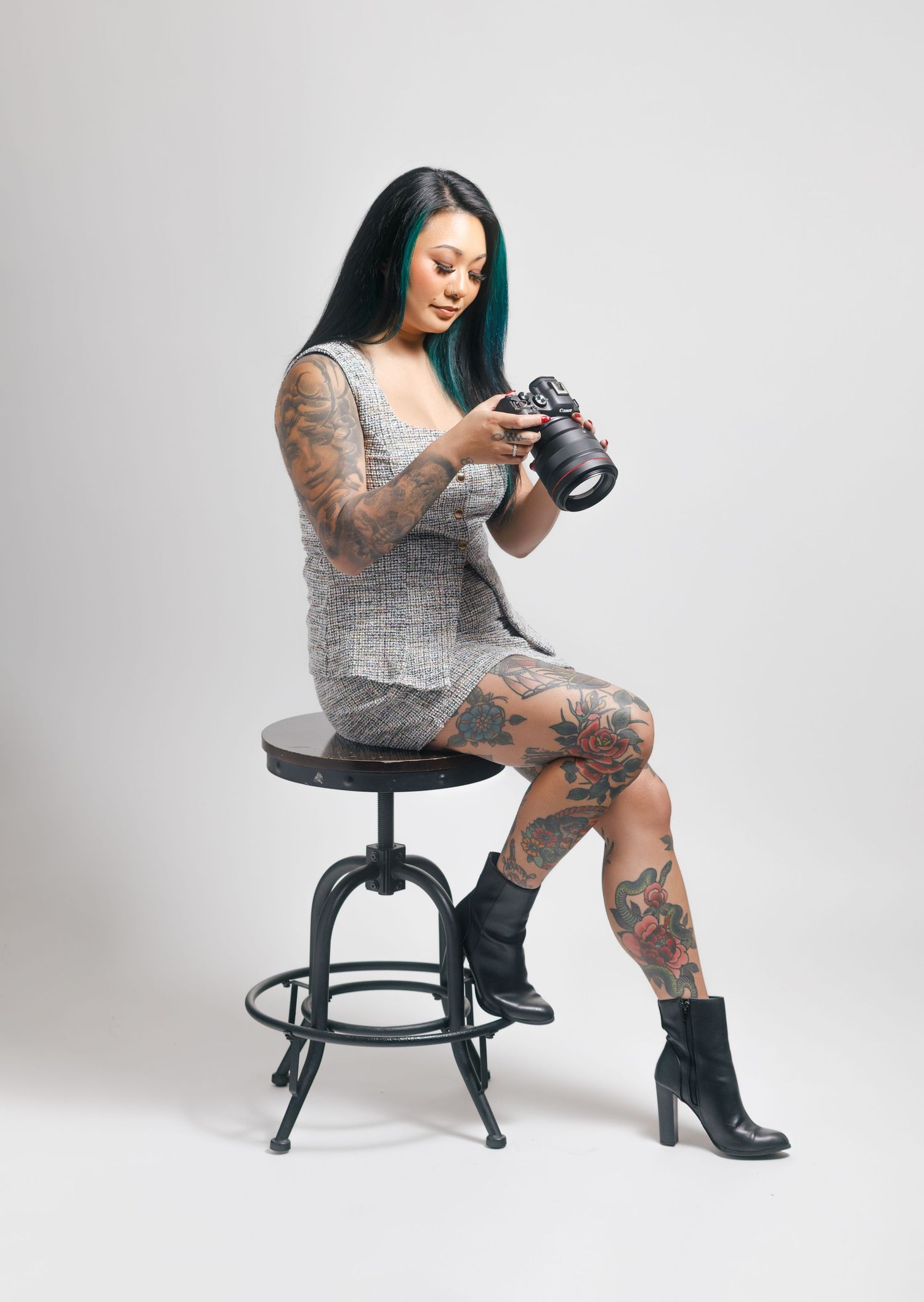 Tattooed woman with teal hair sits on stool reviewing photos on DSLR camera, wearing gray dress and black boots.