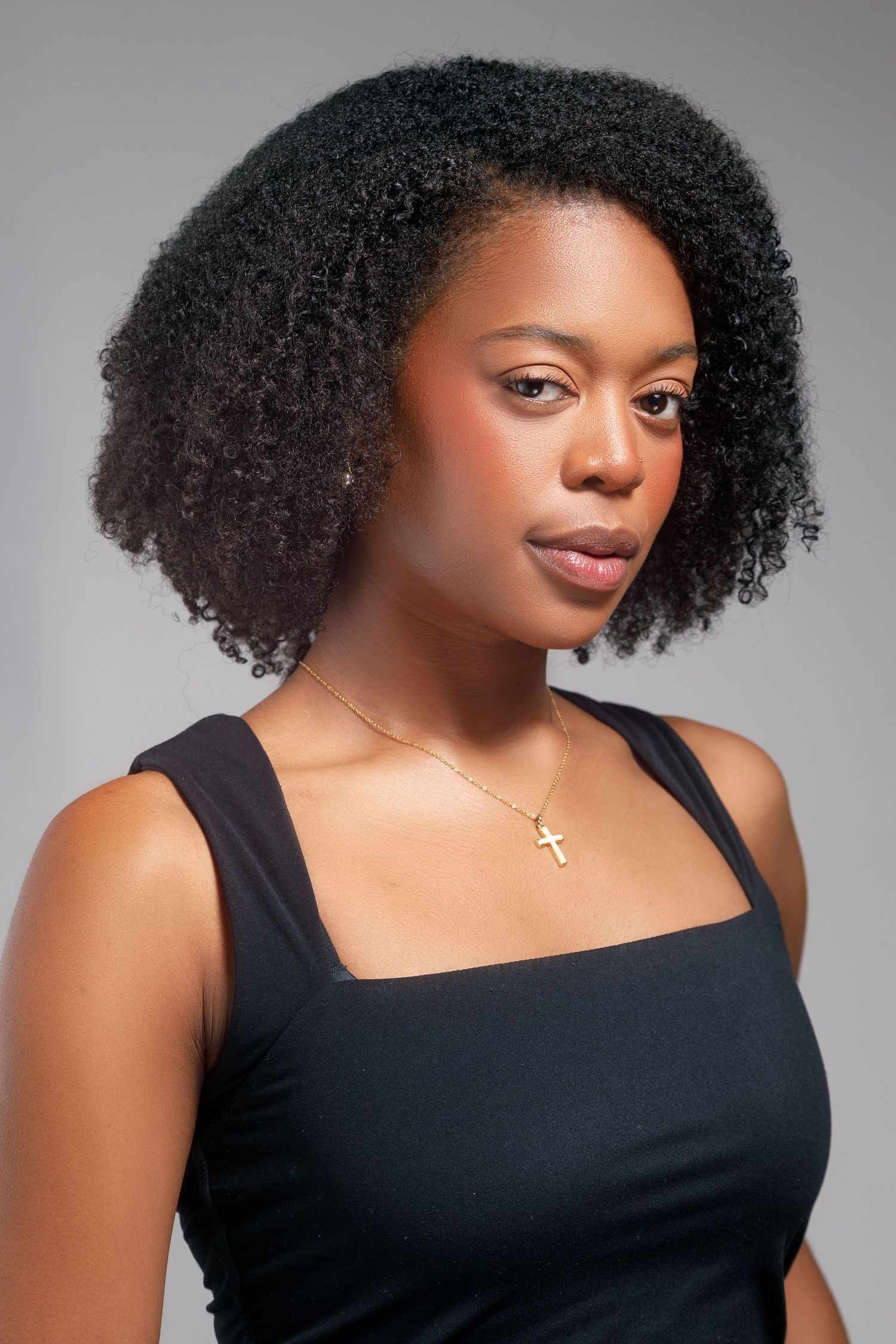 Young Black woman with natural curly hair wearing a black sleeveless top and gold cross necklace against gray background.