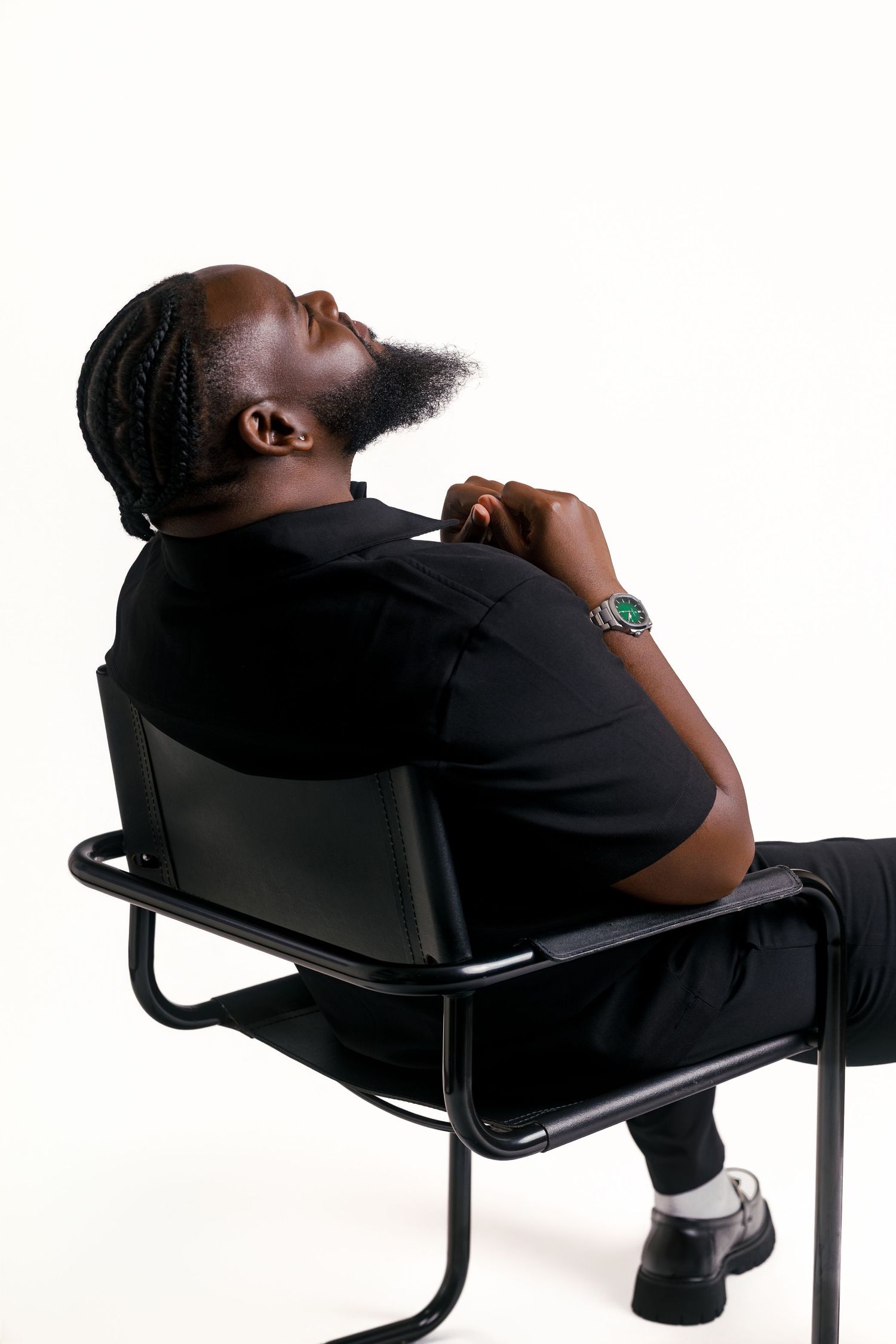 Black man with braids and beard sits on chair, wearing all black outfit and green watch, against white background.