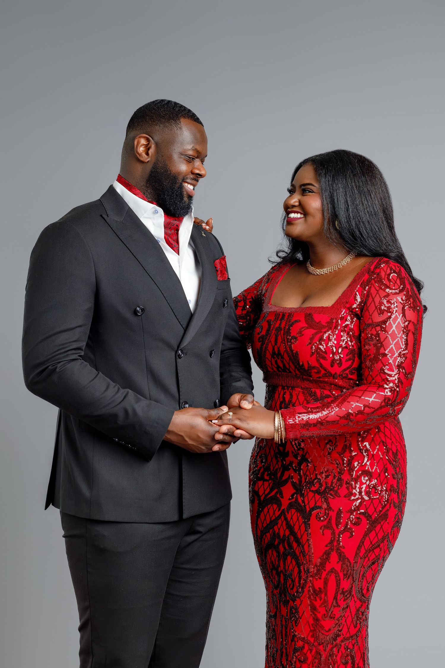 Elegant couple in matching red and black formal attire holding hands and smiling at each other.