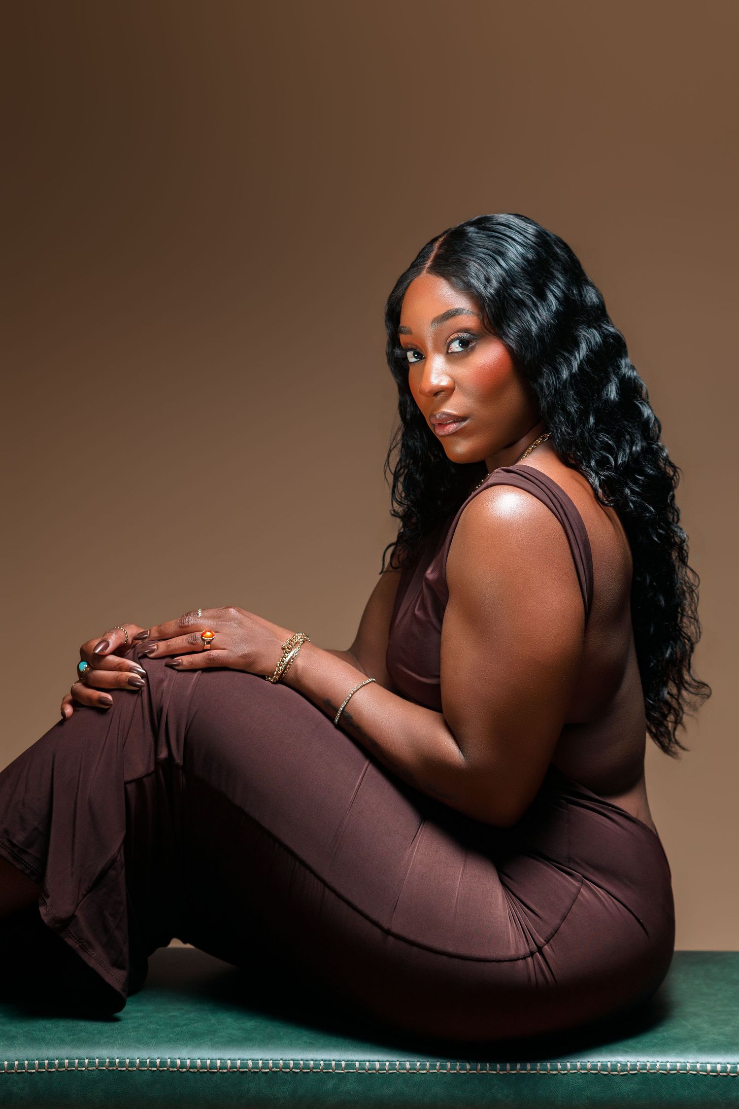A Black woman in a brown outfit sits gracefully on a green surface, wearing gold jewelry, with long wavy hair.