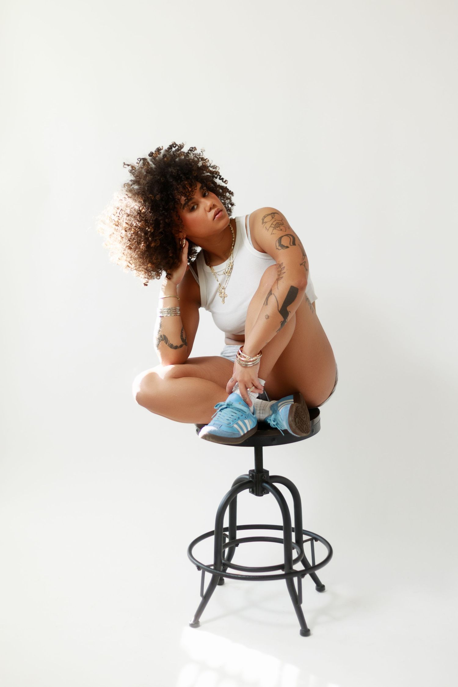 Tattooed woman with curly hair sits cross-legged on black metal stool wearing white bodysuit and blue sneakers against white background.