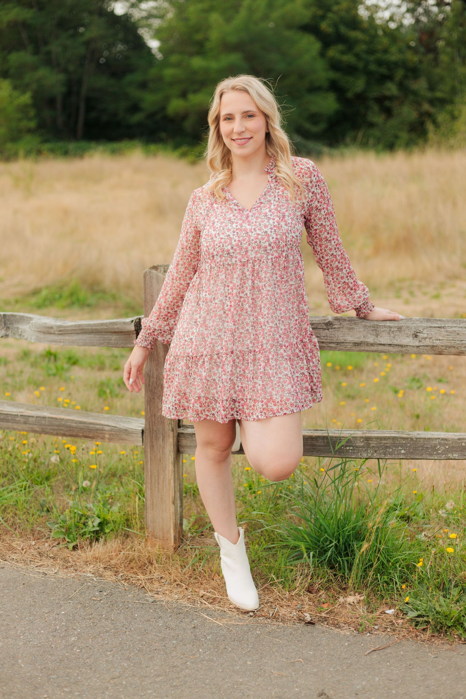 Blonde woman in pink floral mini dress and white boots leaning on wooden fence in a sunny field.