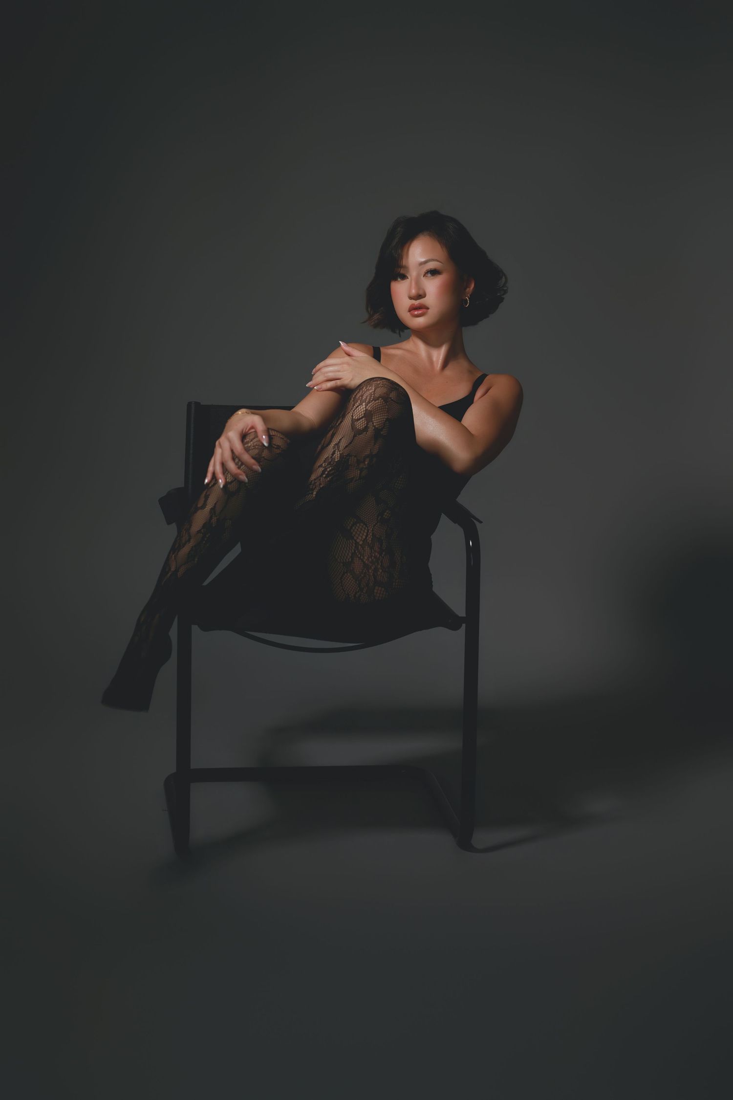 Woman in black lingerie and lace stockings posing elegantly on a chair against a dark gray background.