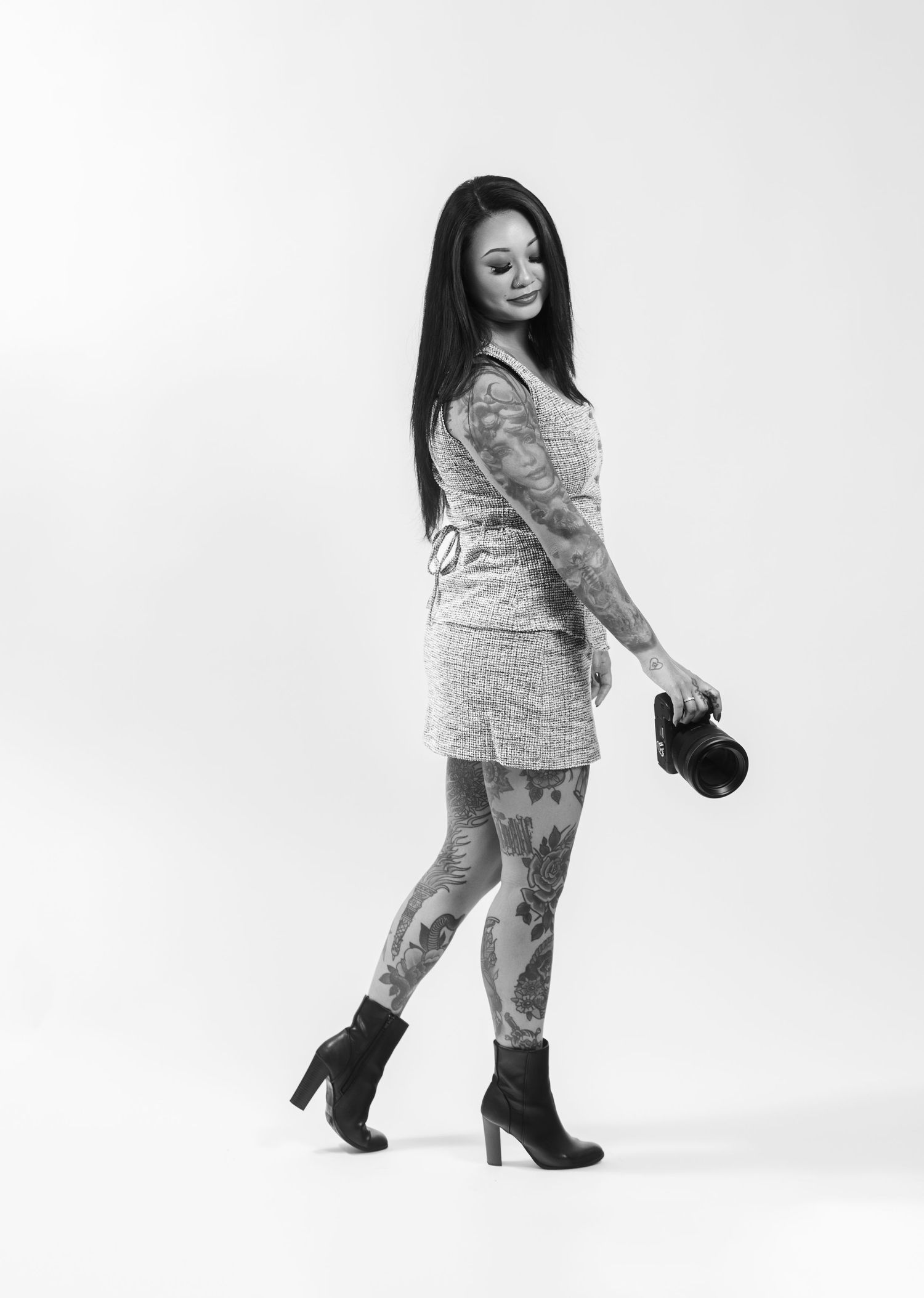 Black and white photo of a tattooed woman in a mini dress and heeled boots holding a camera, smiling over her shoulder.