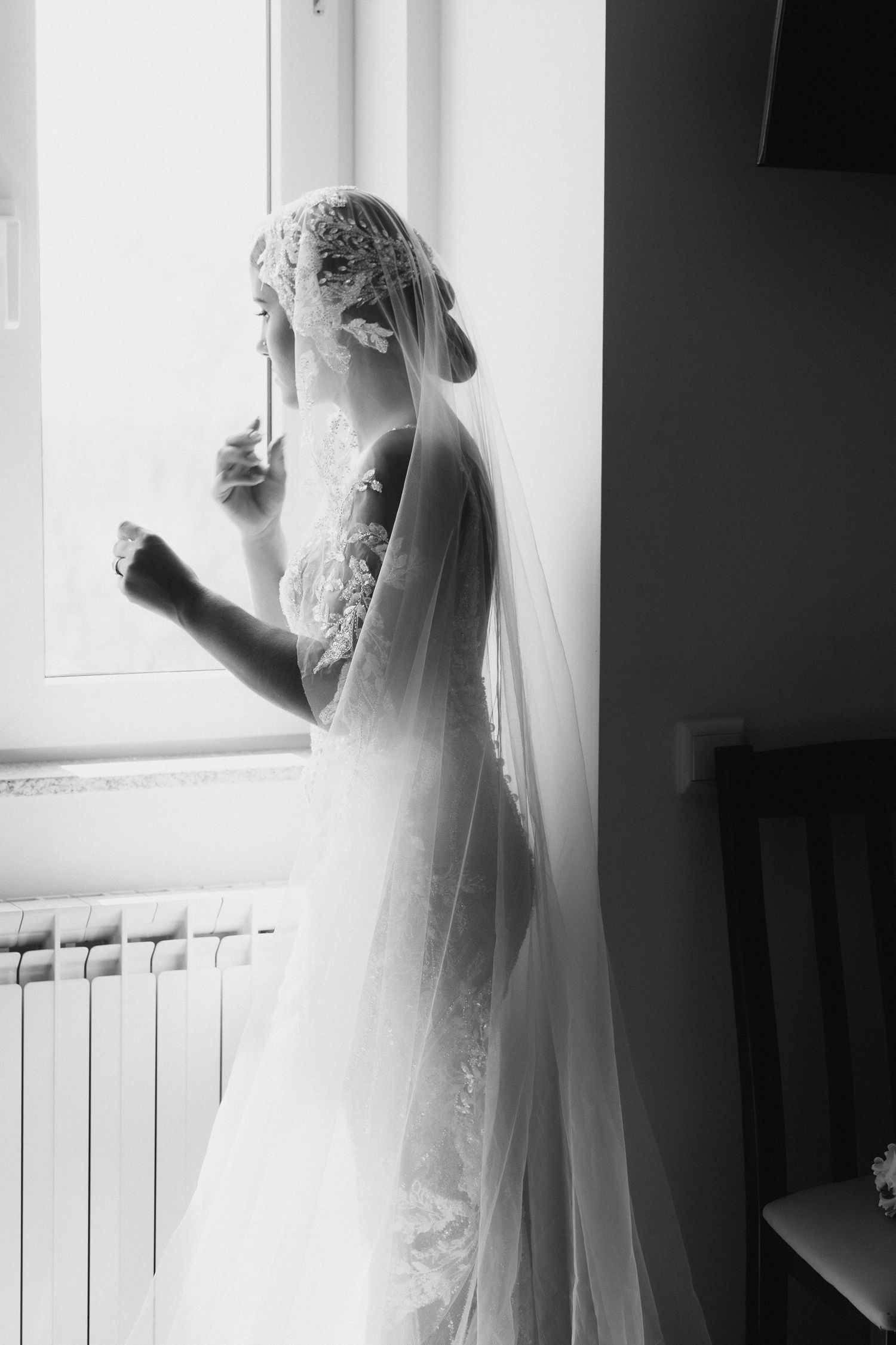 Elegant bride in lace wedding dress and veil stands by window in black and white portrait photo.