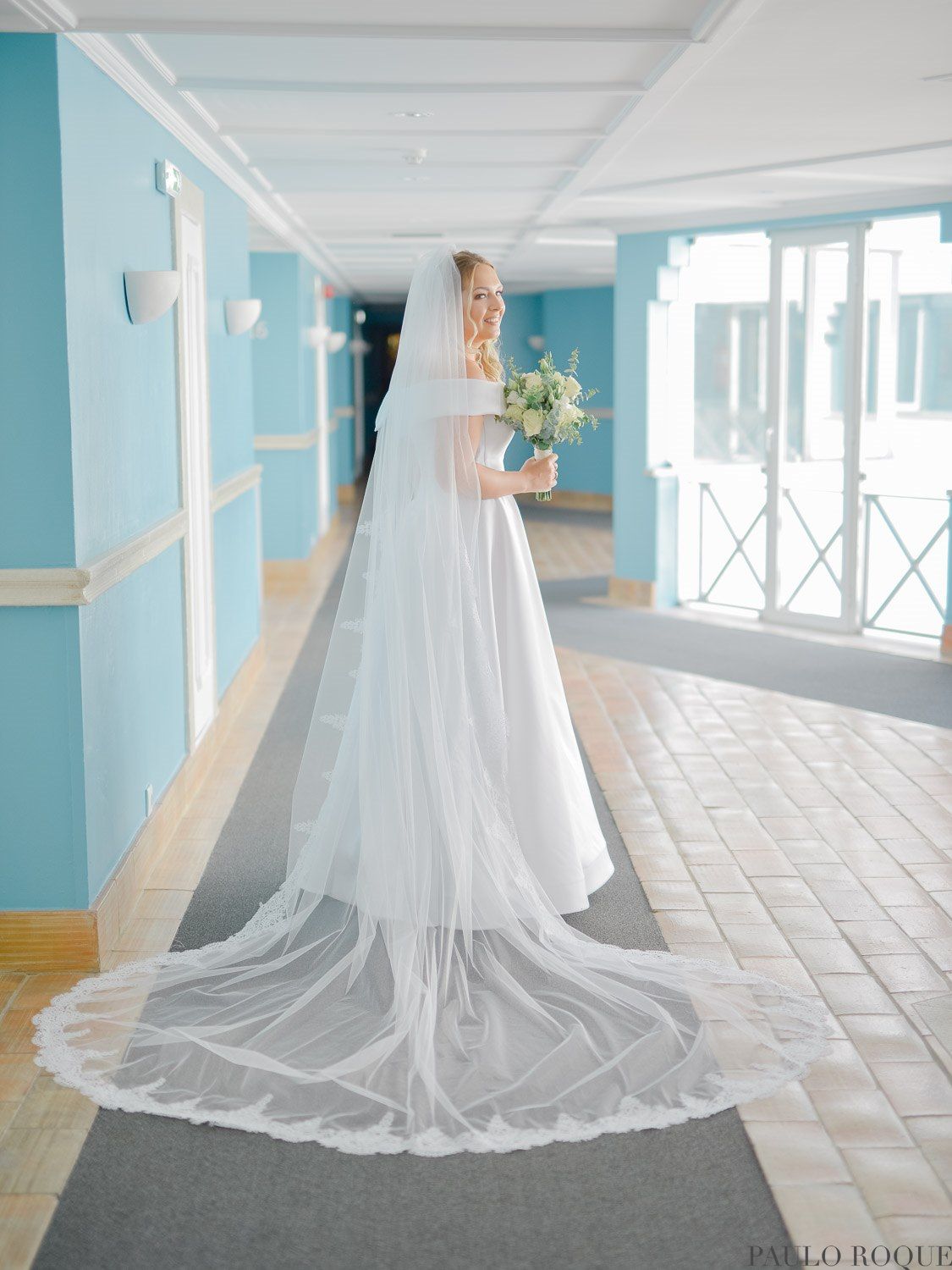 Noiva de vestido branco elegante e longo véu catedral, segurando um buquê em um corredor azul, casamento em Evora.