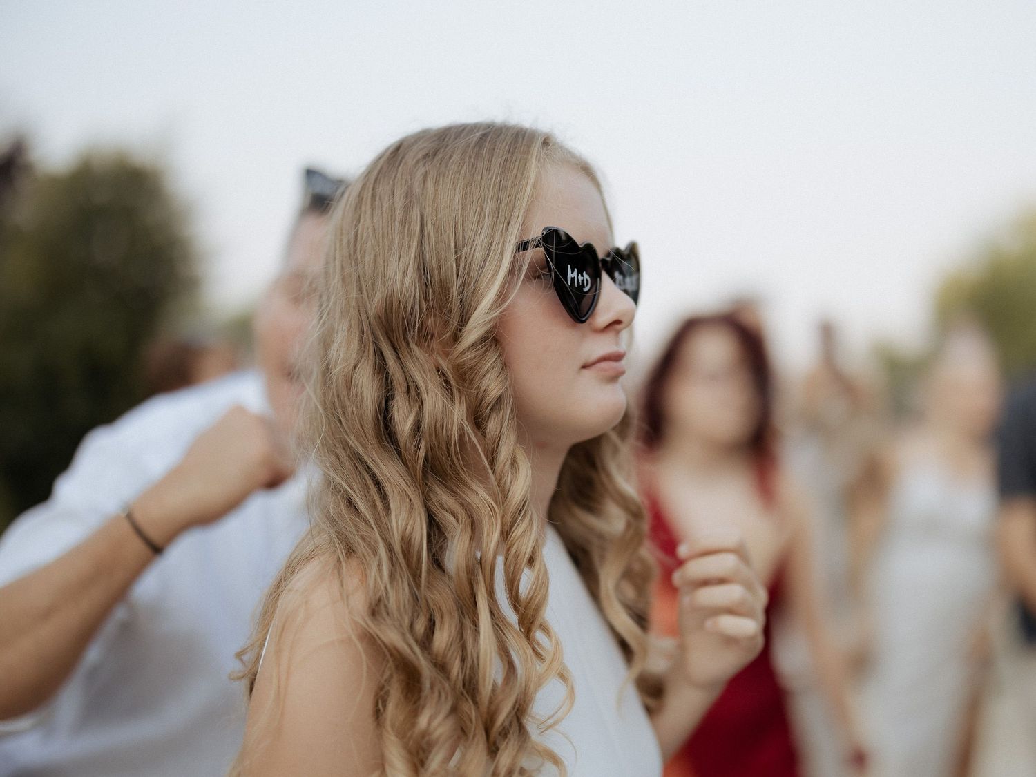 Young blonde woman wearing black sunglasses at an outdoor event with blurred crowd in background.