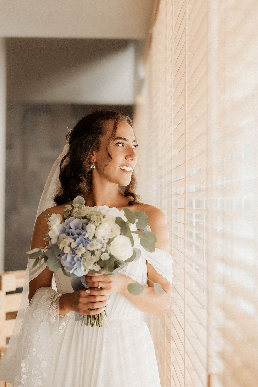 Noiva sorridente de vestido branco a segurar um buquê de flores azuis e brancas e usando véu, de pé junto a uma janela em Vila do Conde.