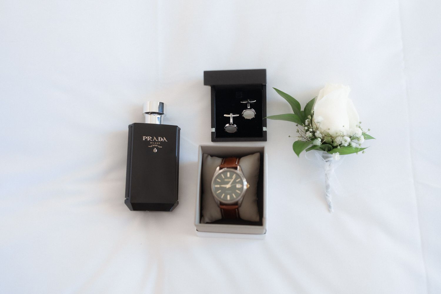 Perfume Prada, abotoaduras em caixa, relógio de couro marrom e flor de lapela branca dispostas sobre tecido branco para um casamento.