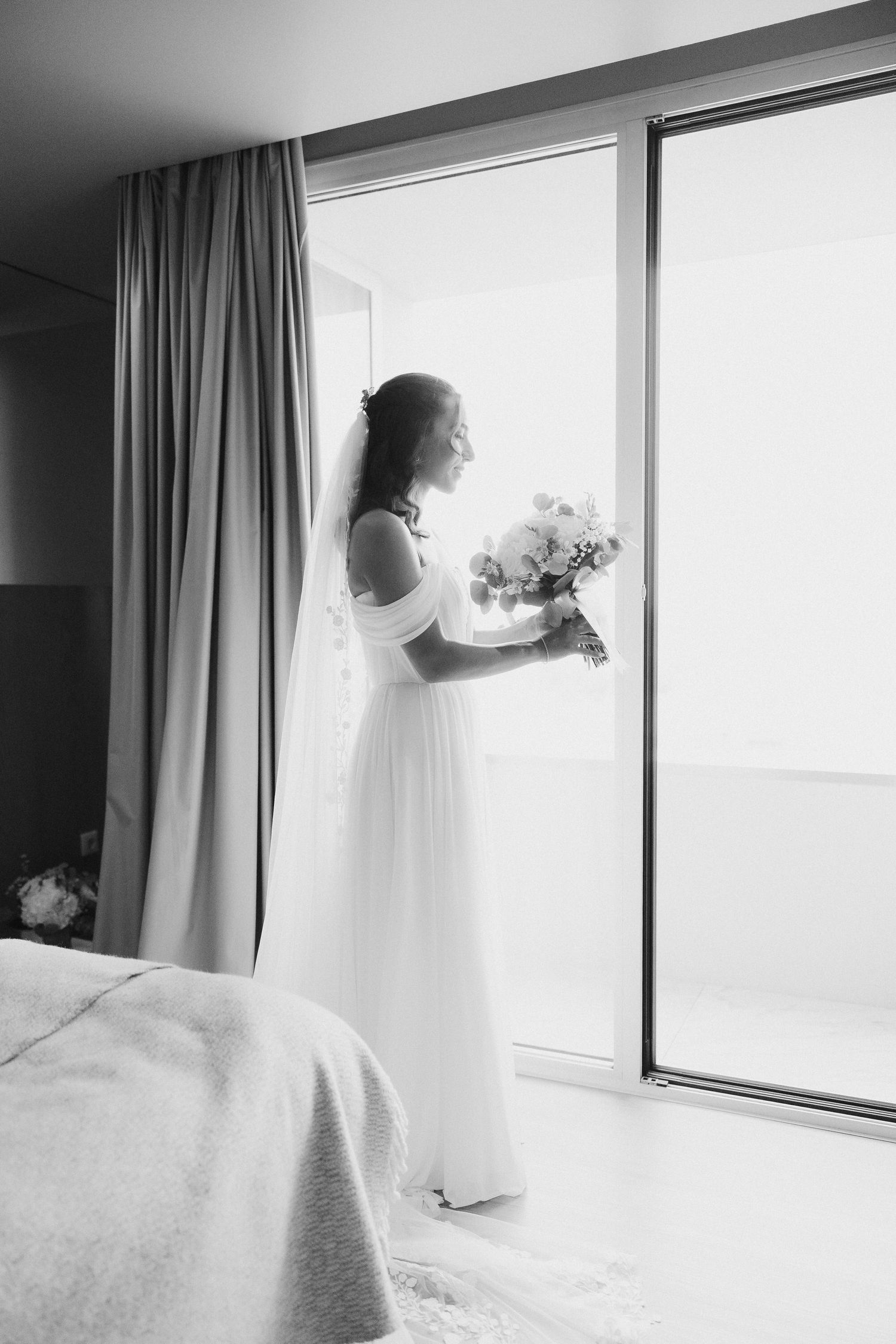 Noiva de vestido branco e véu segura buquê junto a uma grande janela; foto de casamento em preto e branco. Casamento no Set Lounge Azurara,