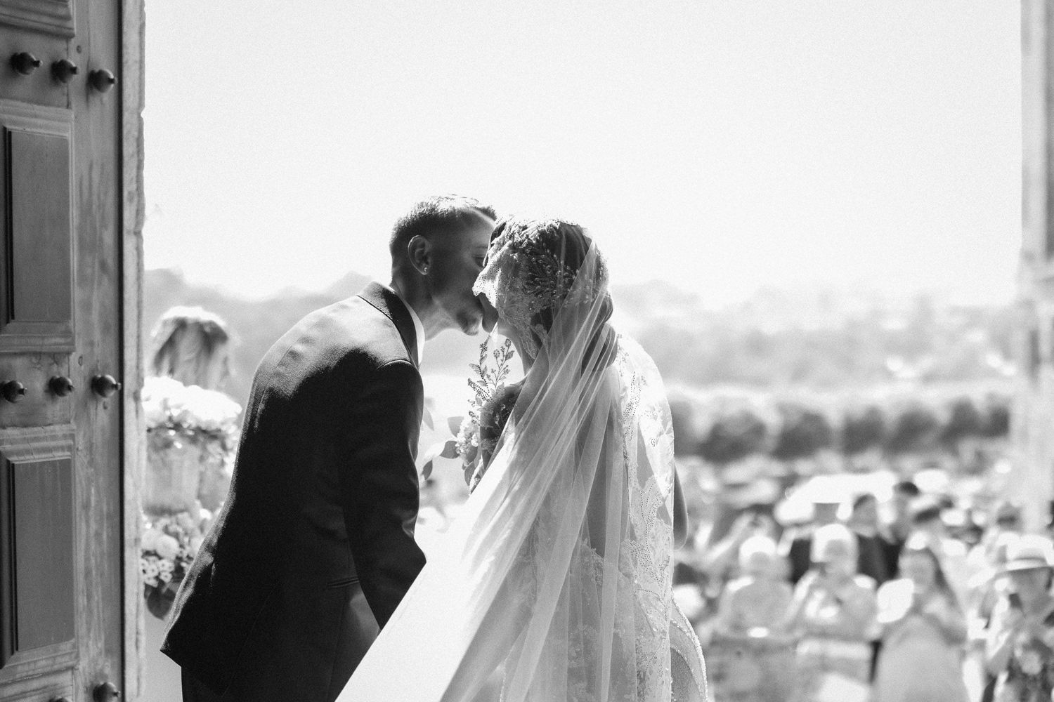 Black and white photo of a bride and groom sharing their first kiss at a sunny outdoor wedding ceremony.