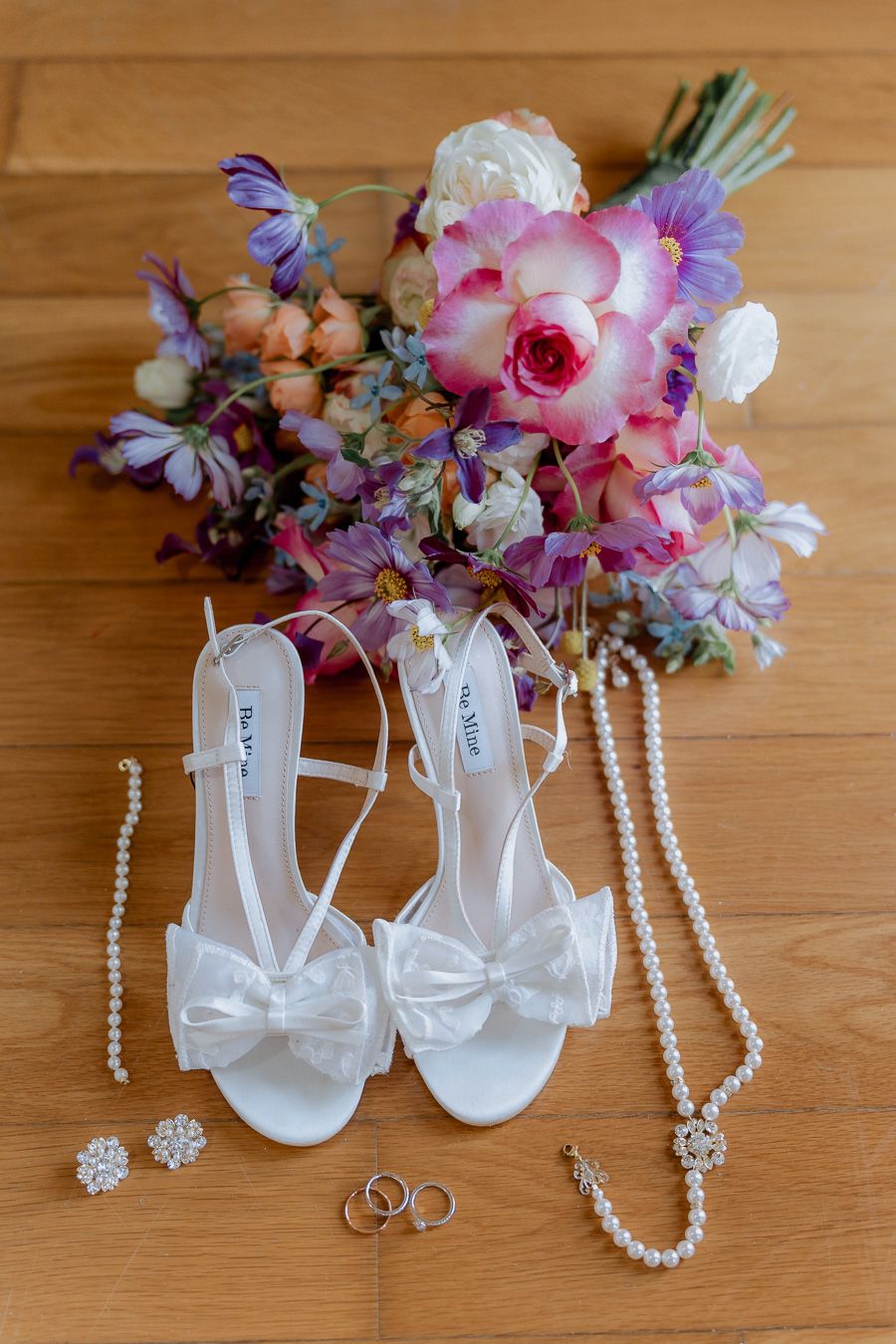 Sapatos de noiva brancos com laços ao lado de joias de pérolas, anéis de ouro e um buquê de flores. Casamento Viana do Castelo