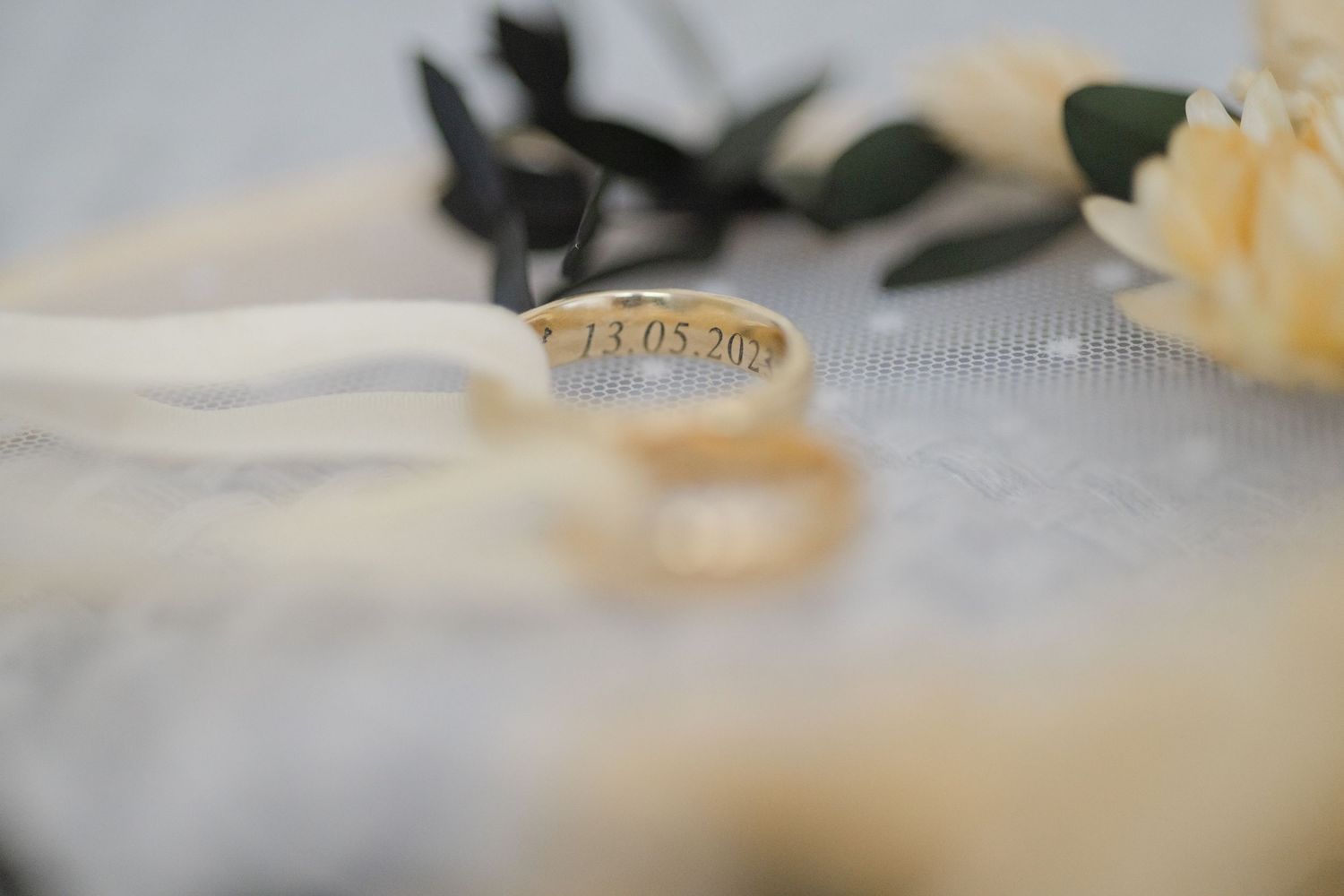 Aliança de casamento de ouro com a data 13.05.20 gravada, posicionada junto a flores brancas e uma fita sobre tecido de renda.