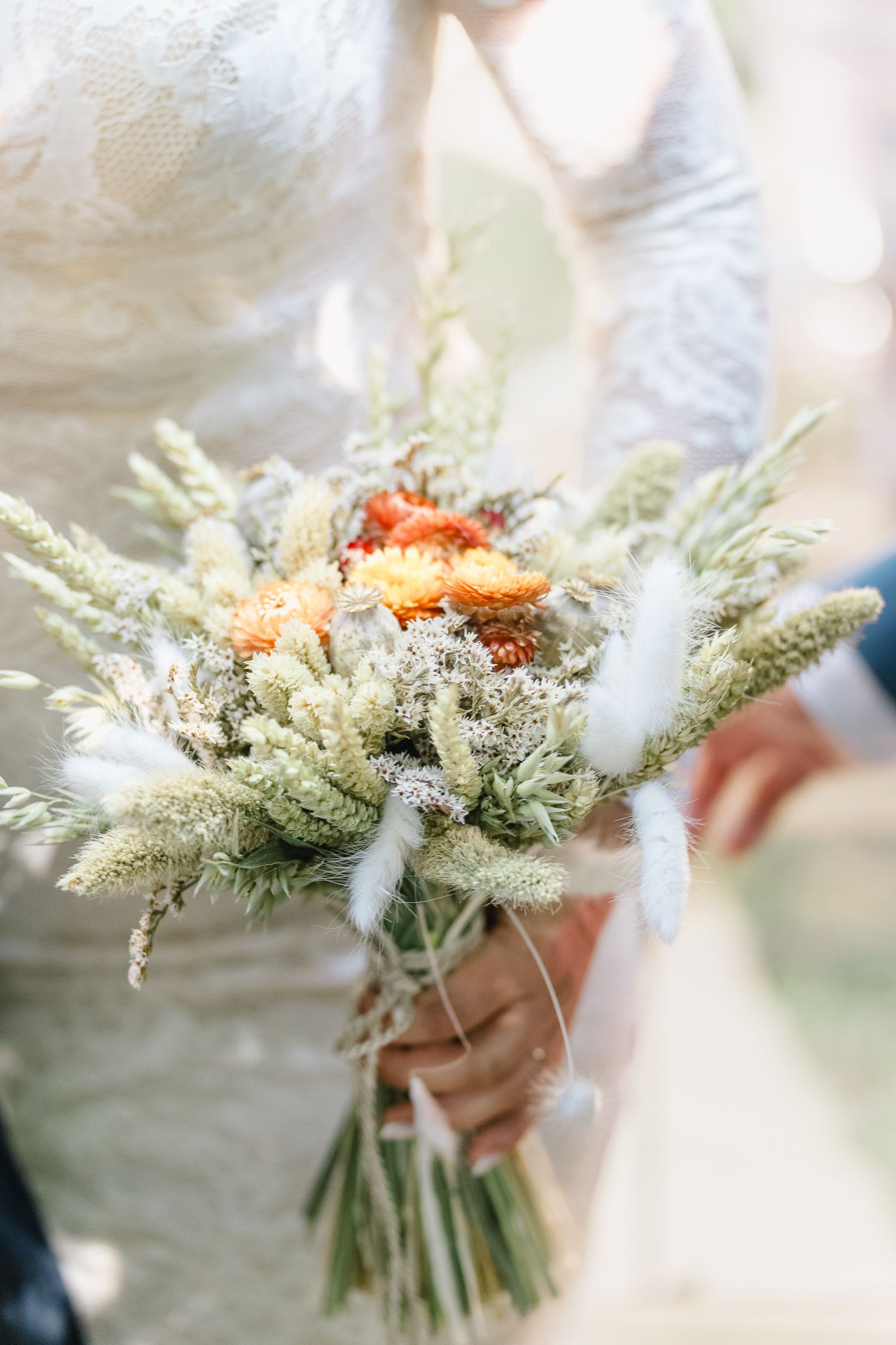 Noiva de vestido de renda segurando um buquê de casamento estilo boho rústico com gramíneas secas, penas brancas e flores laranjas.