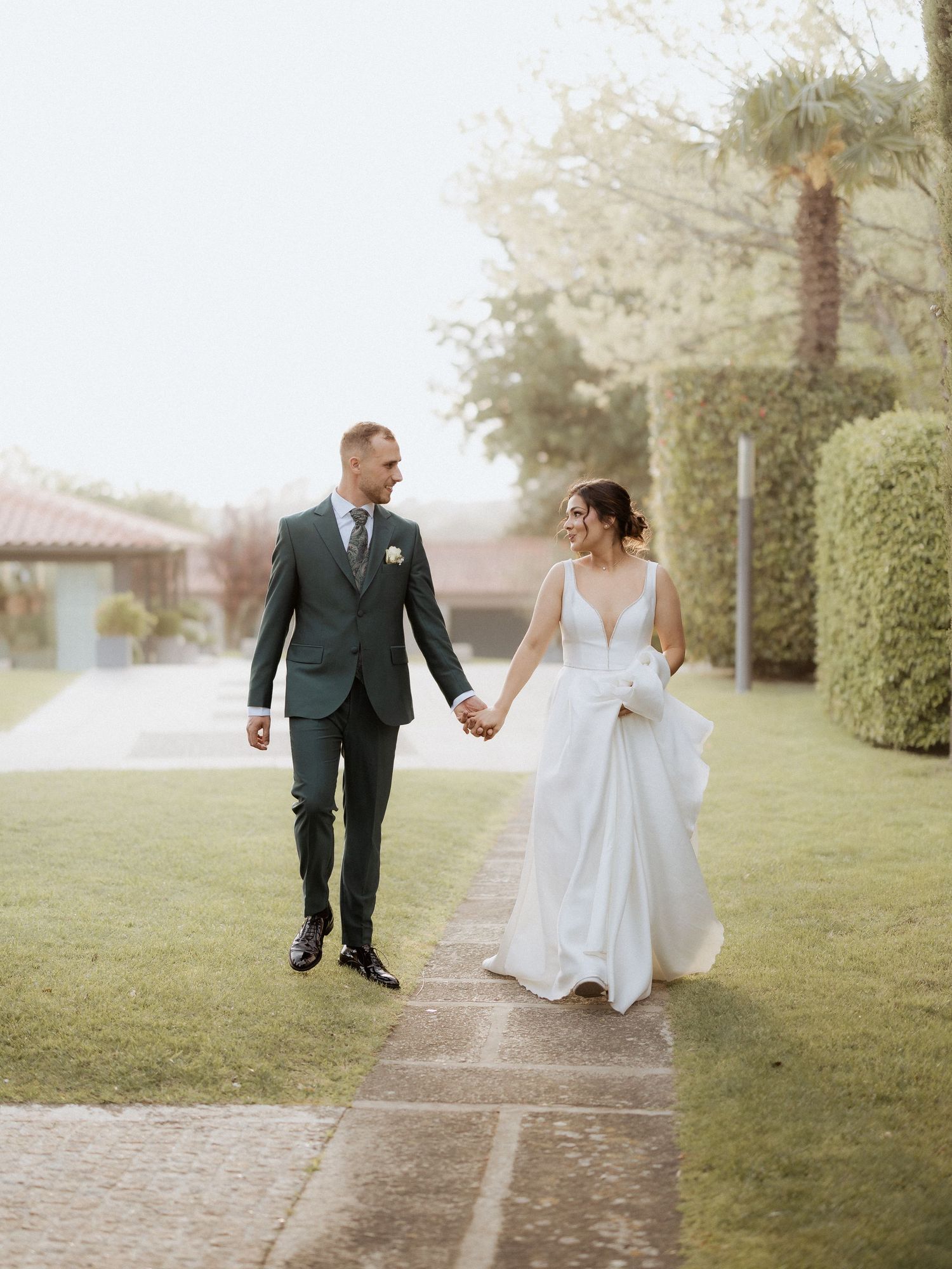 Uma noiva de vestido branco e um noivo de fato escuro caminham de mãos dadas por um jardim no dia do casamento na Quinta de Santo André.