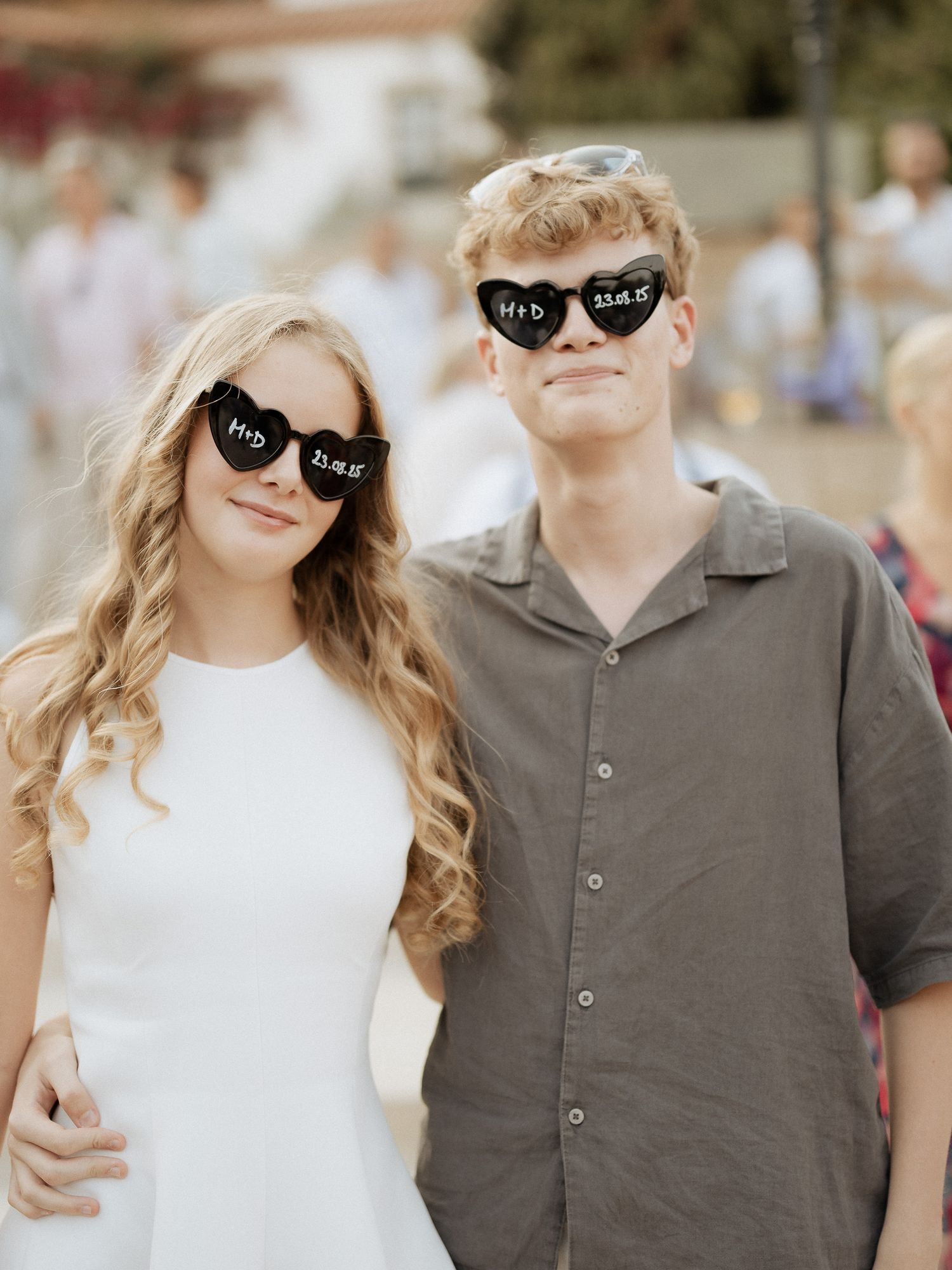 Casal jovem usando óculos de sol pretos iguais em formato de coração com texto em branco, posando junto ao ar livre em um evento ensolarado.
