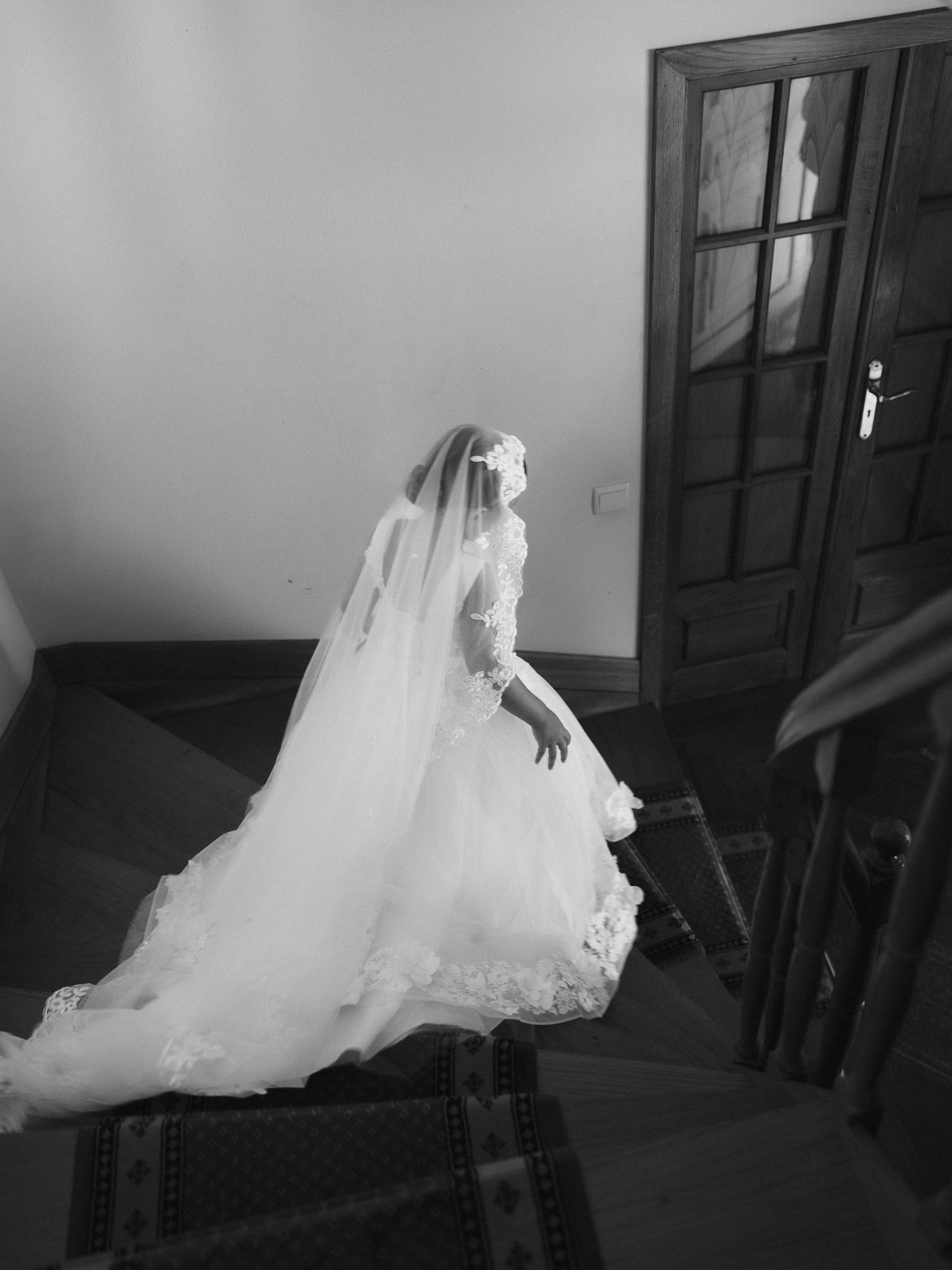 Foto em preto e branco de uma noiva com um vestido de casamento fluido e véu, sentada em uma escada perto de uma porta de madeira.