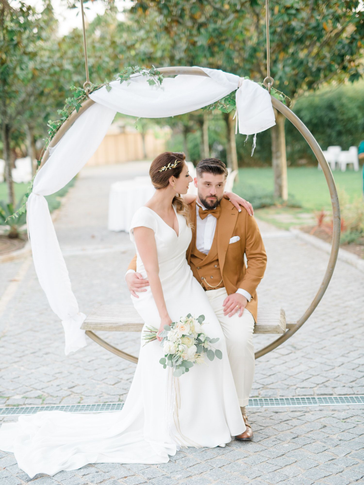 Os noivos trocam um beijo em um balanço circular decorado com cortinas brancas e folhagens, ao ar livre na Quinta do Carvalho.