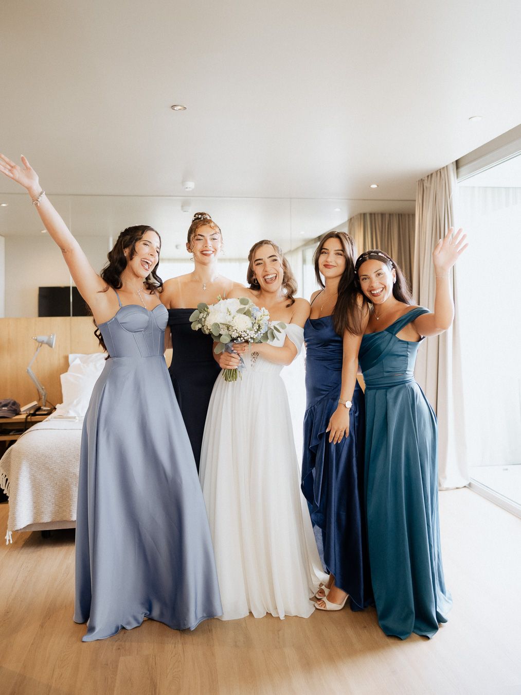 A noiva, segurando o buquê, posa com três damas de honra vestidas de azul, sorrindo e acenando. Casamento Set Lounge Azurara.