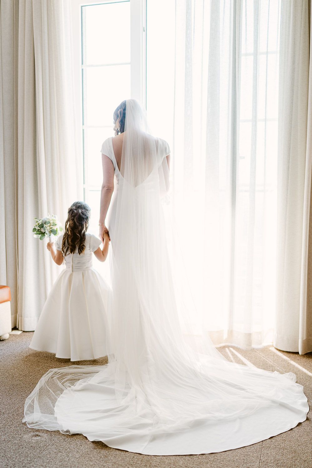 A noiva, vestida de branco dama de honra, também de branco, ambas  diante de uma janela bem iluminada. casamento na Pousada de Santa Luzia