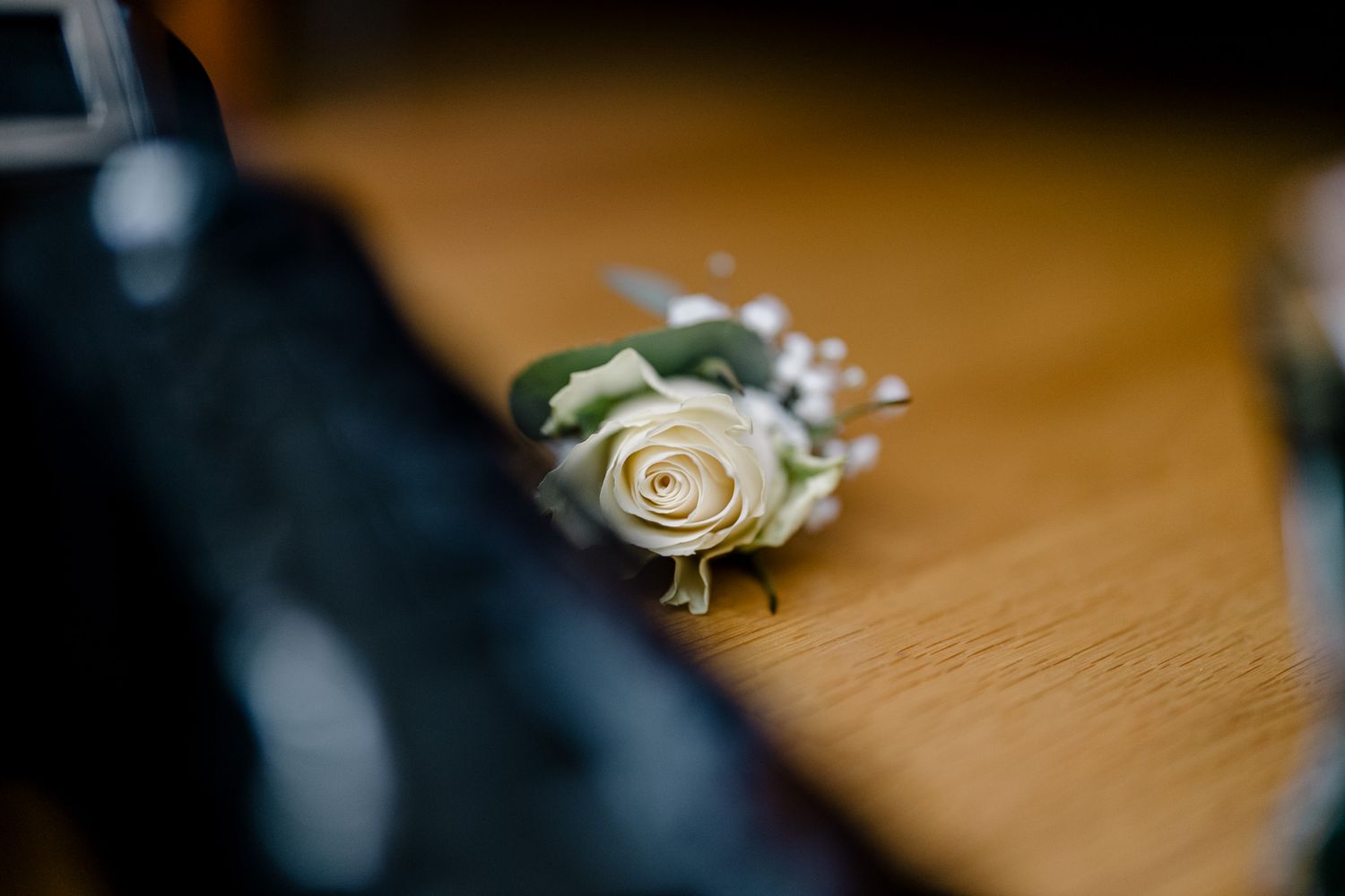 Uma delicada flor branca na lapela, adornada com folhagens, repousa sobre uma superfície de madeira ao lado de sapatos escuros de casamento.
