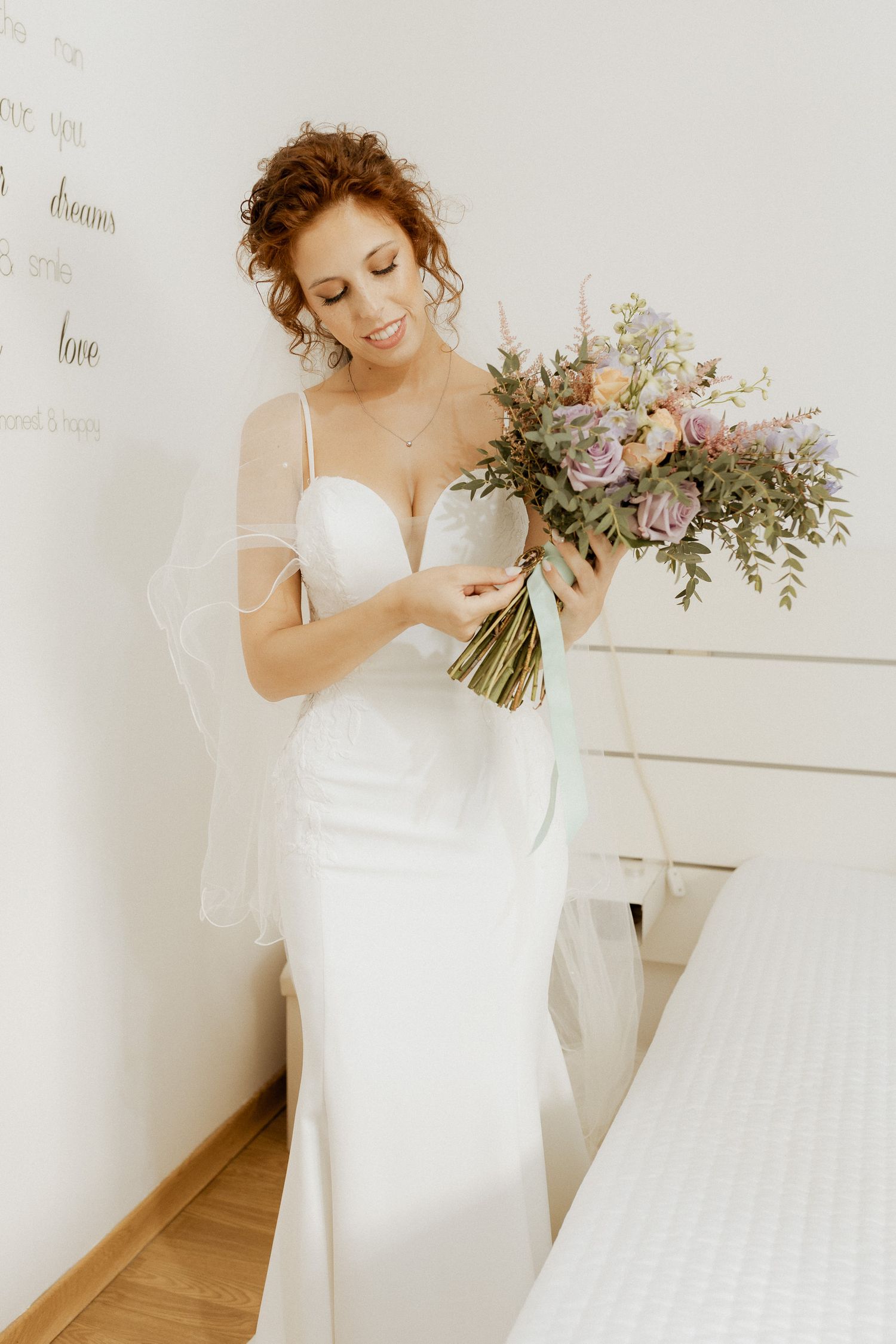 Noiva de vestido de cetim branco segura um buquê de flores em tons pastel, com rosas e folhagens, sorrindo suavemente em um ambiente branco