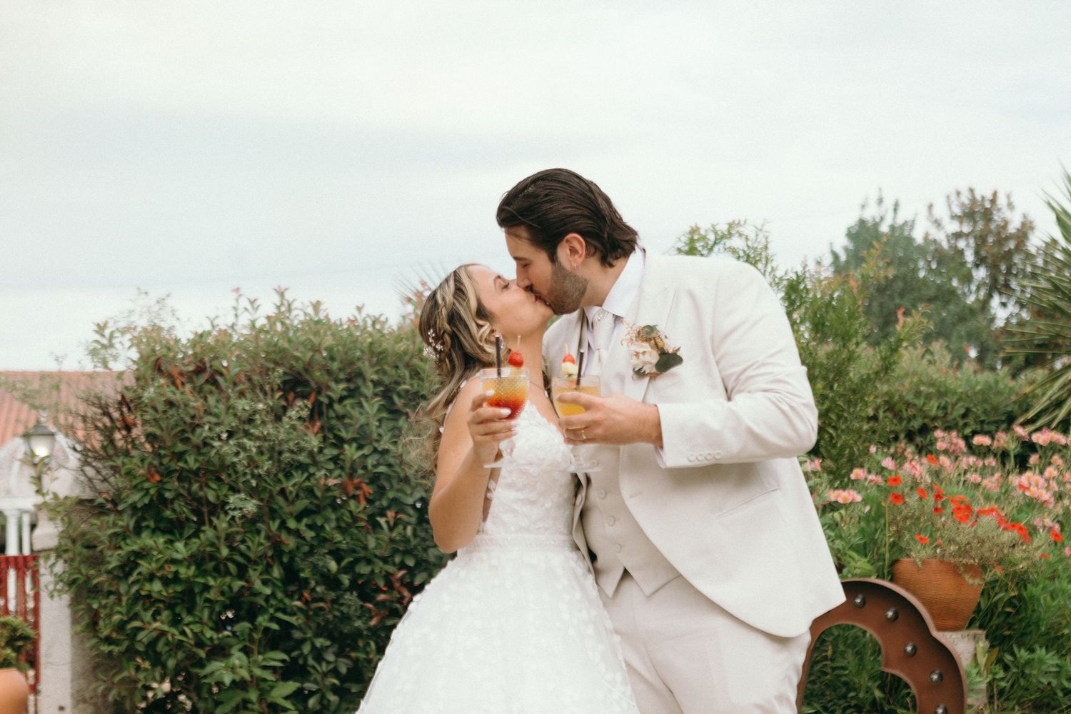 Casal recém-casado trocam um beijo enquanto seguram taças de champanhe ao ar livre, durante a comemoração do casamento. Quinta do Cruzeiro.