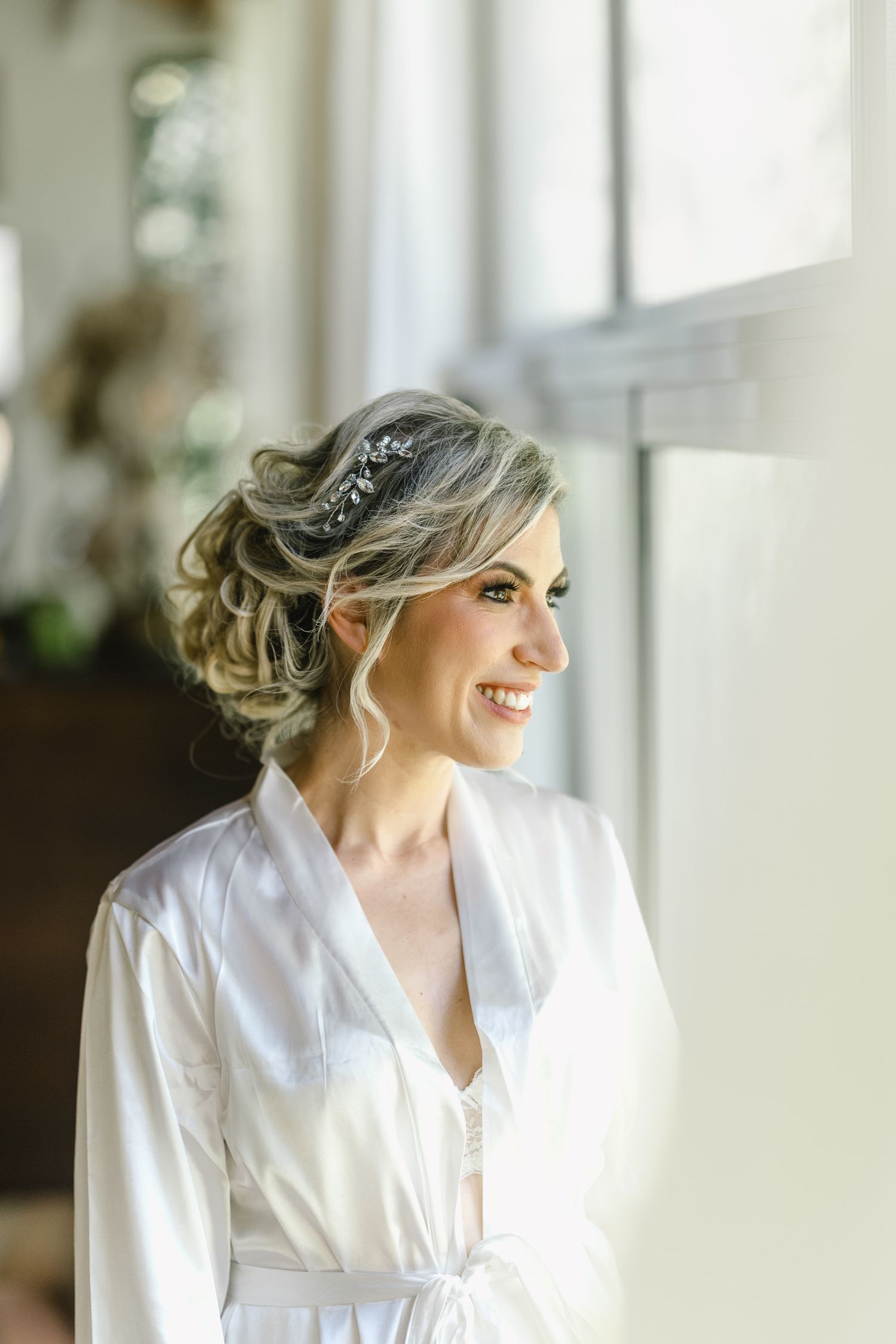 Noiva sorridente em vestido de cetim branco, com um penteado preso elegante e acessório floral no cabelo, de pé junto a uma janela iluminada