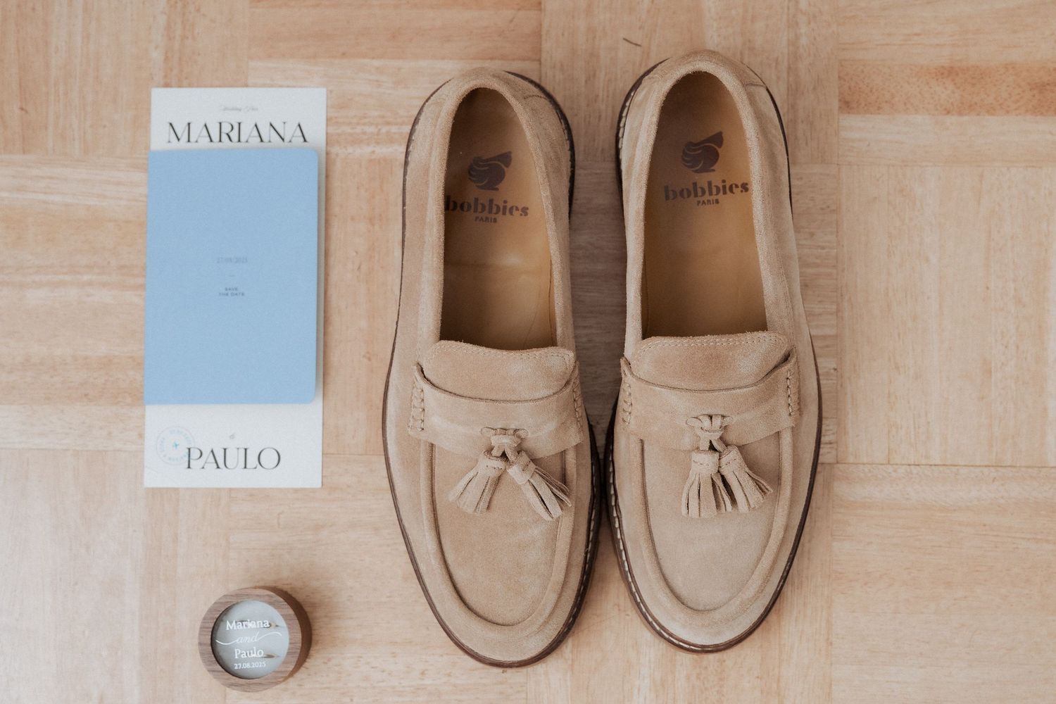 Mocassins de camurça bege com borlas expostos no chão de madeira ao lado dos convites de casamento de Mariana e Paulo. Set Lounge Azurara