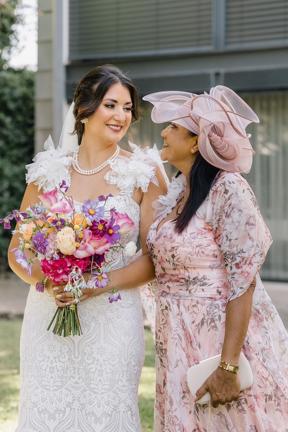Noiva em vestido de renda com detalhes em penas segura buquê colorido ao lado da mãe. Casamento na Quinta do cruzeiro