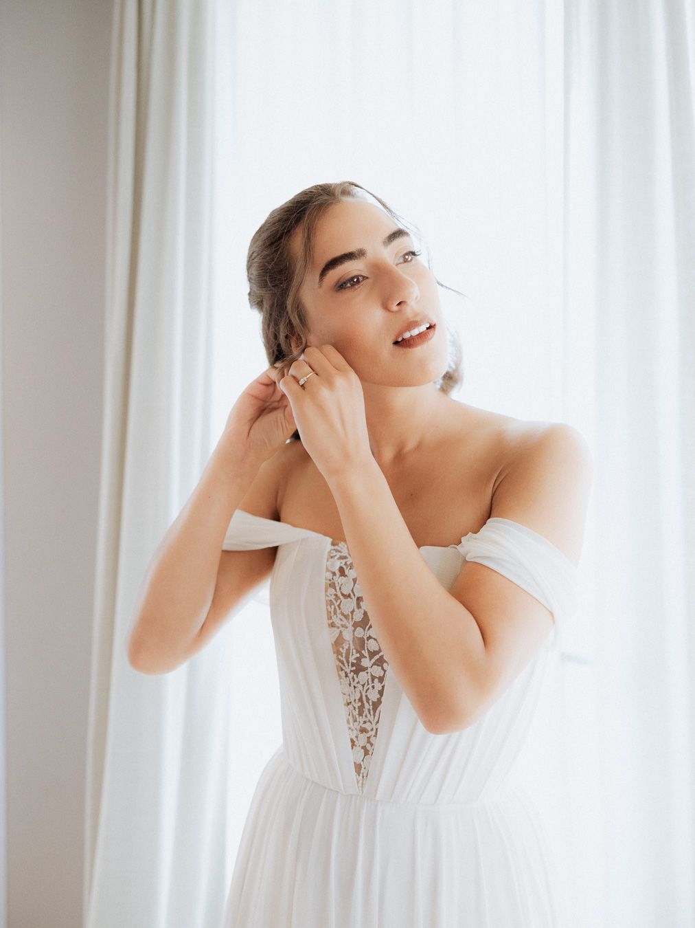 Noiva em vestido de noiva branco, colocando brincos perto de uma janela bem iluminada, casamento na Quinta Santo André Barcelos