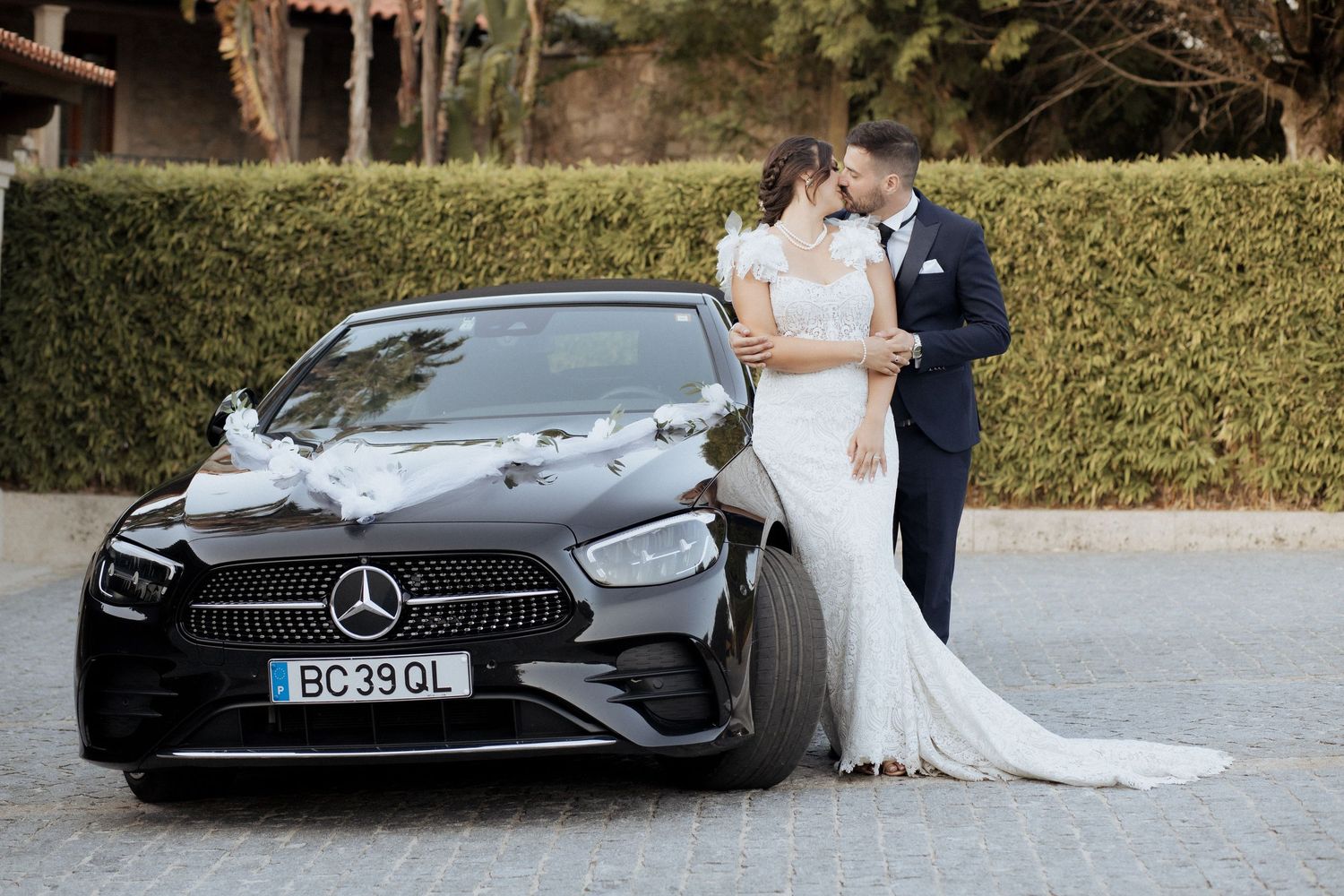 Noivos se beijando ao lado de um carro de casamento Mercedes-Benz preto decorado, ao ar livre. Casamento na Quinta do Cruzeiro.