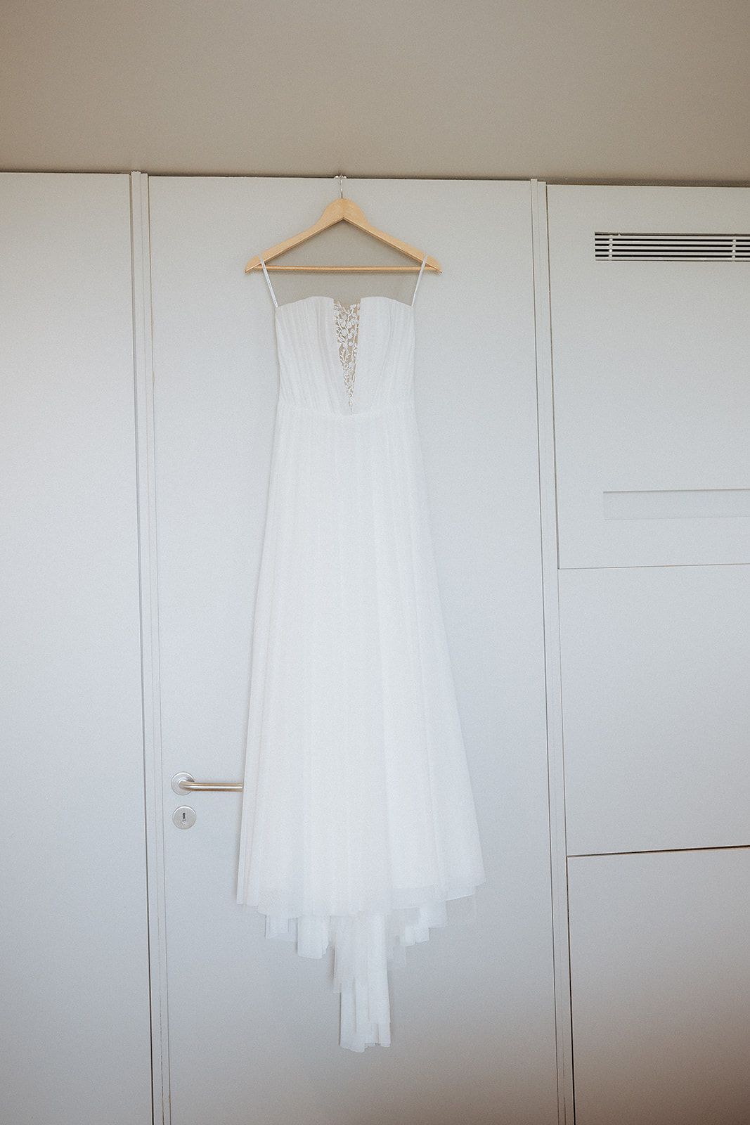 Vestido de noiva branco e elegante pendurado em um cabide de madeira, contra uma parede branca e um guarda-roupa.