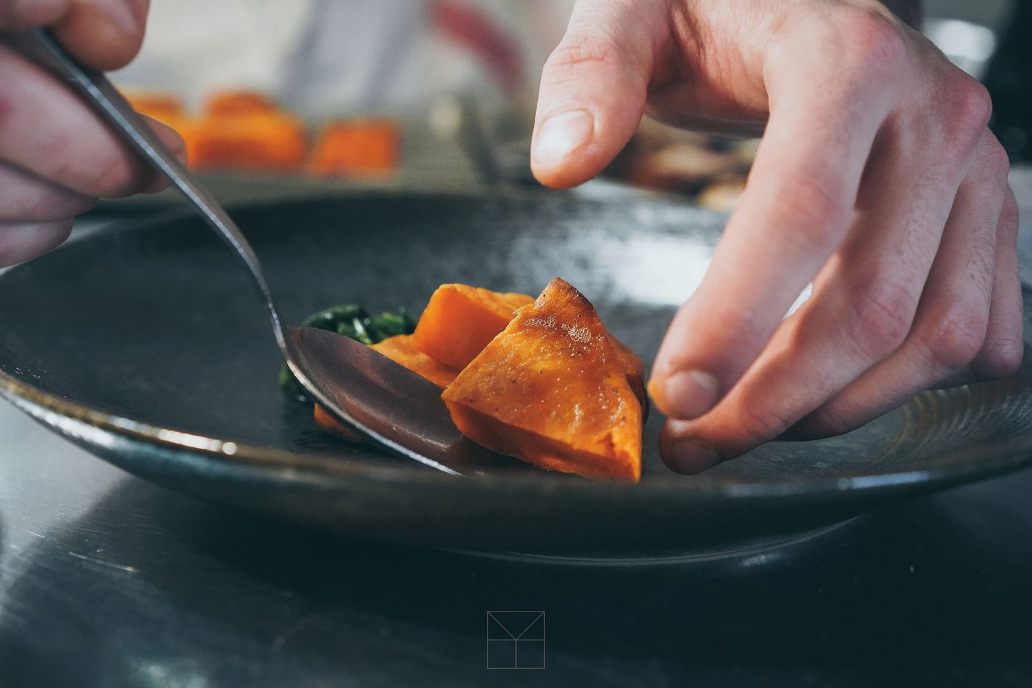 Chef plating an orange glazed dish on a dark ceramic plate in a professional kitchen.