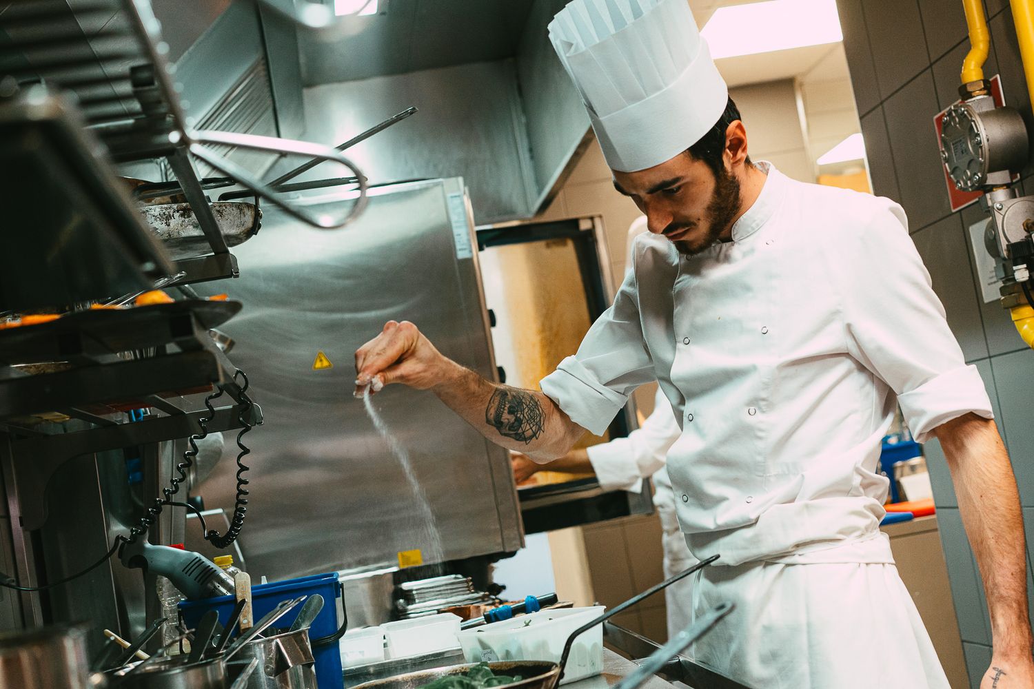 A tattooed chef in white uniform and tall hat adds finishing touches to a dish in a restaurant kitchen.
