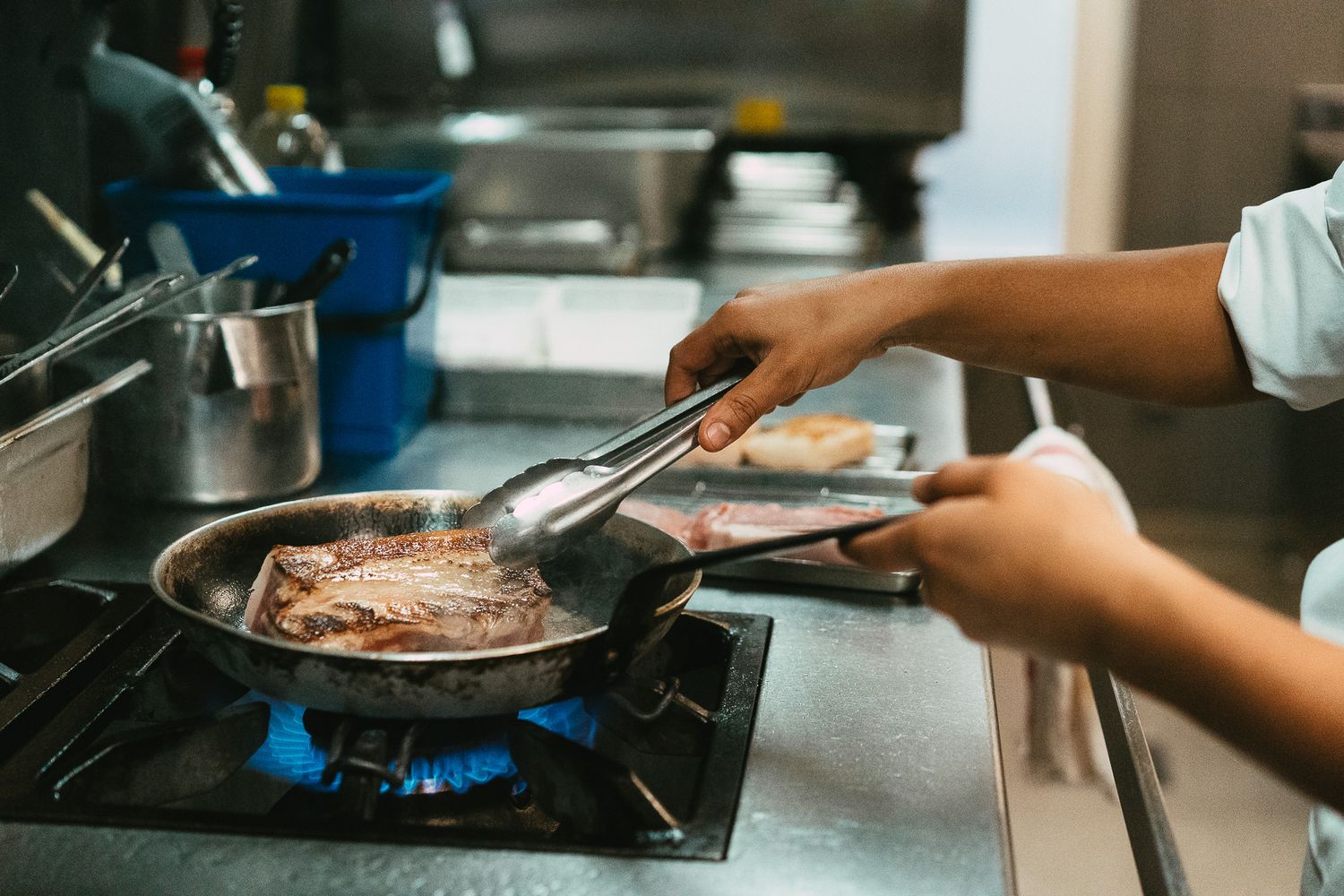Chef using tongs to cook a seasoned steak in a frying pan over a blue gas flame.