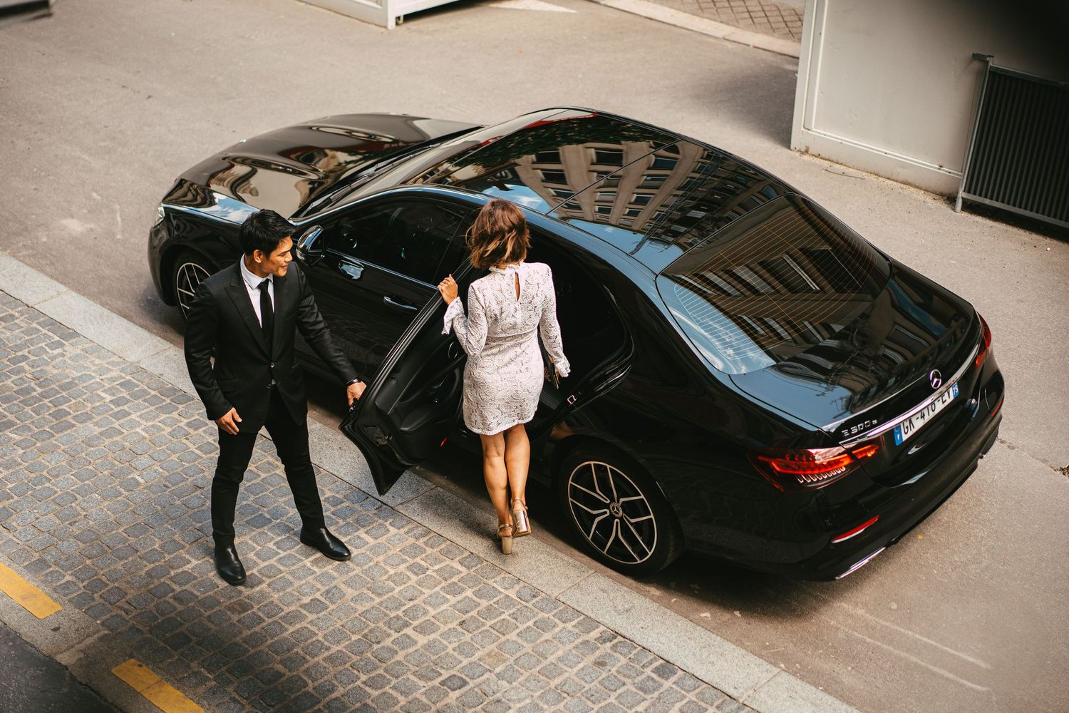 Aerial view of chauffeur escorting elegant woman to black luxury sedan on cobblestone street.