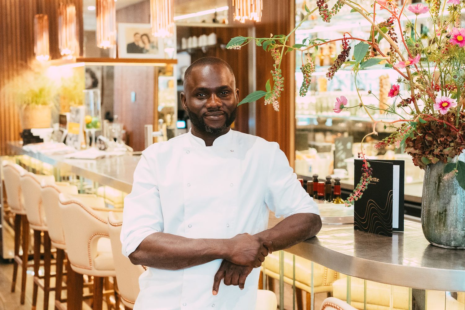 Confident male chef in white uniform with arms crossed posing in a stylish restaurant with floral decor.