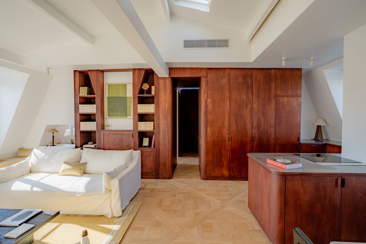 Spacious attic room with rich wooden wardrobes, built-in shelves, skylight, and white sofa seating.