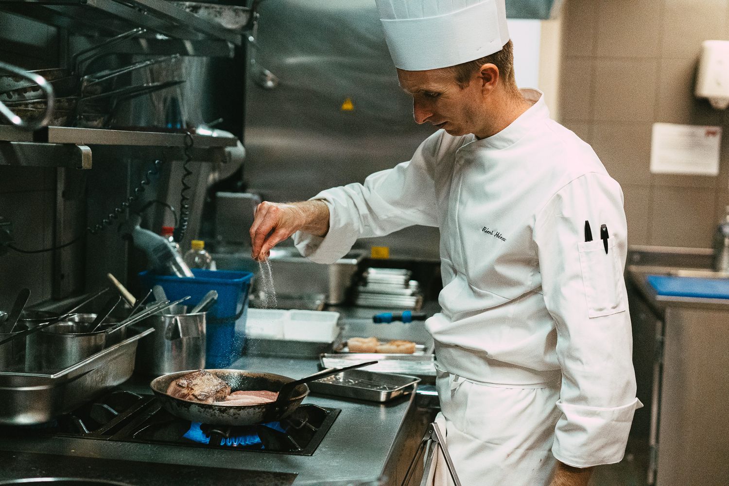 A chef in white uniform seasoning meat in a pan on a gas stove in a professional kitchen.