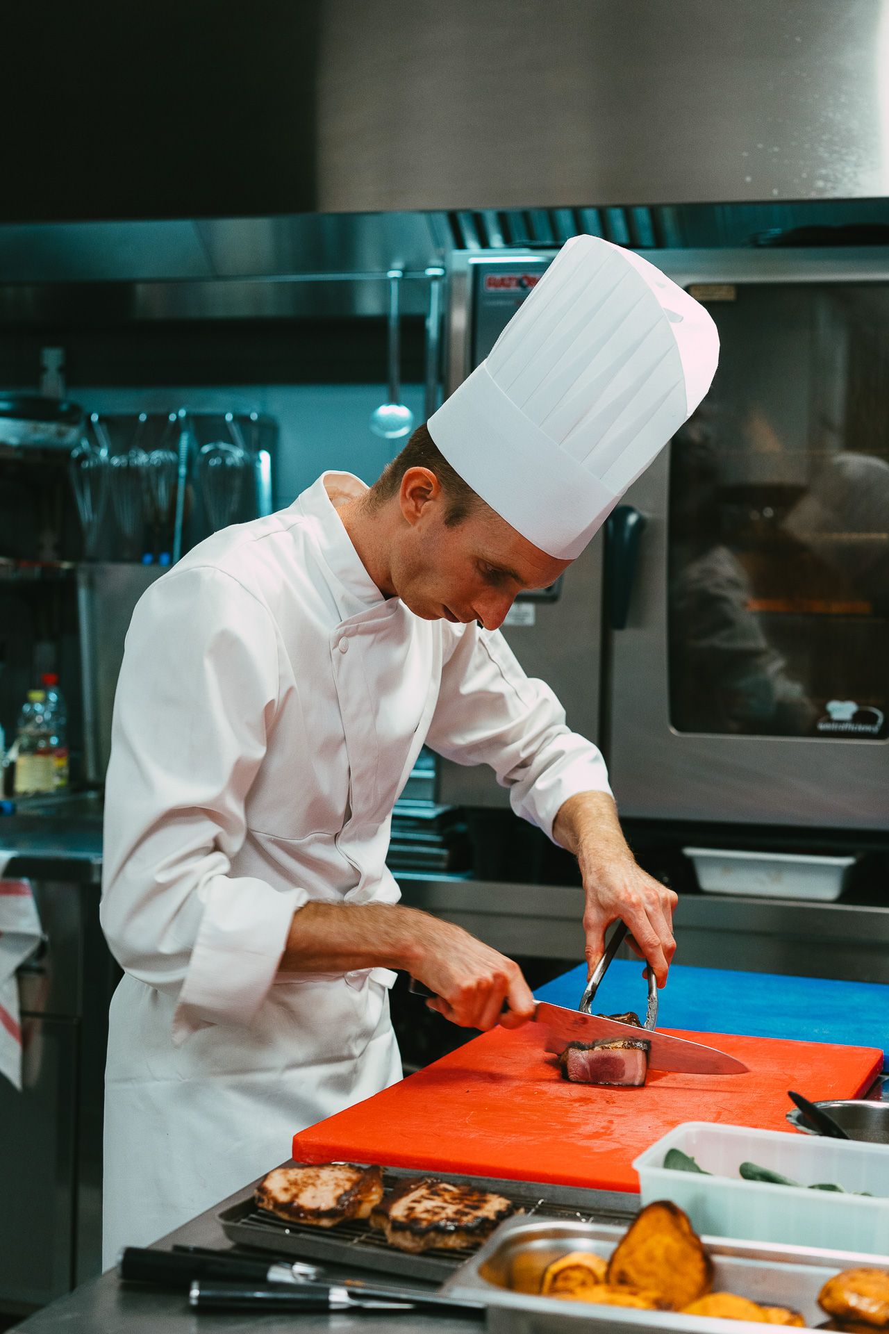 A focused chef in white uniform carefully cuts meat on a red cutting board in a professional kitchen.