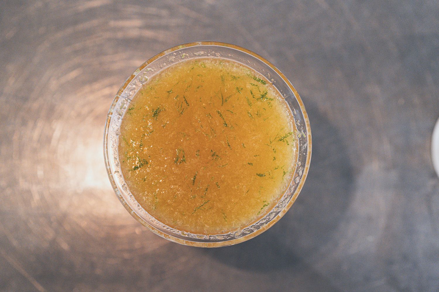 Overhead view of a golden herb-infused liquid in a glass container on a metallic surface.