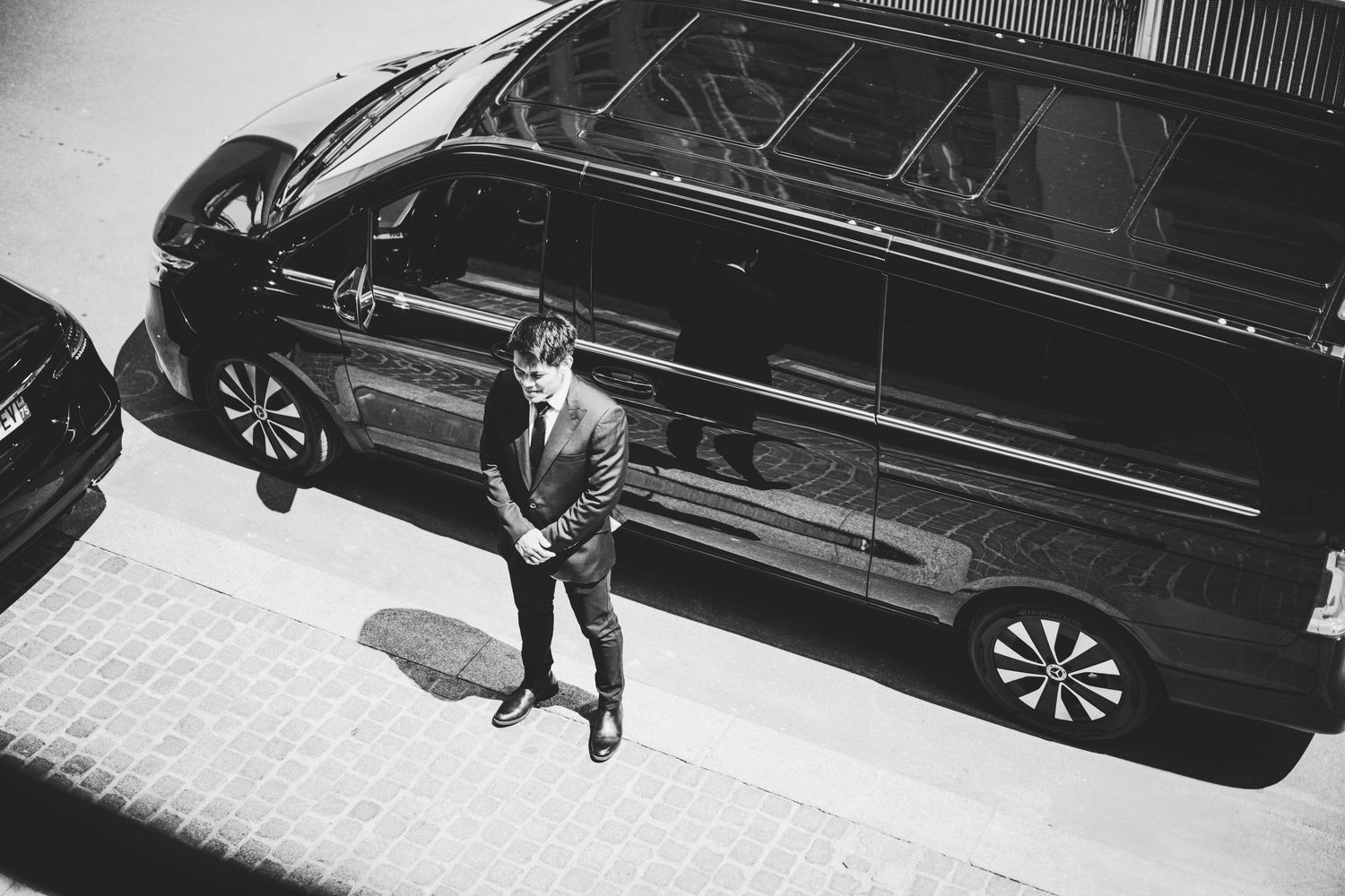Man in suit standing beside a black luxury van on cobblestone street, aerial black and white photo.