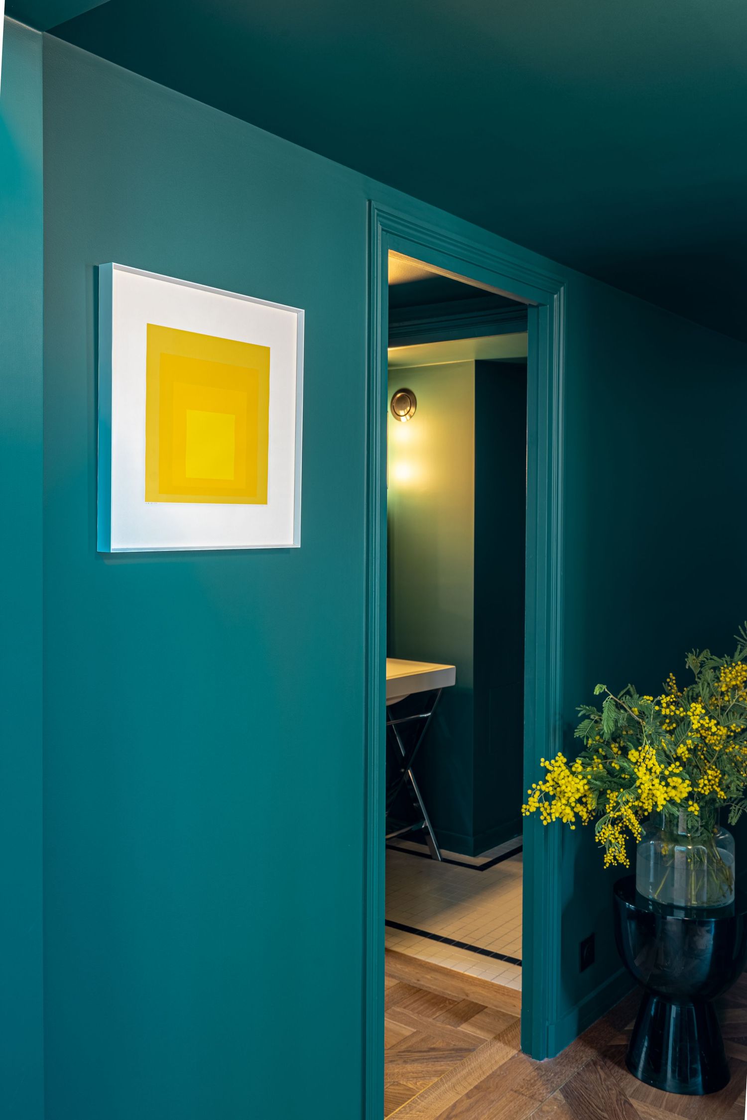 Bold teal-painted hallway with yellow framed artwork, decorative vase, and glimpse into a dark moody study room.